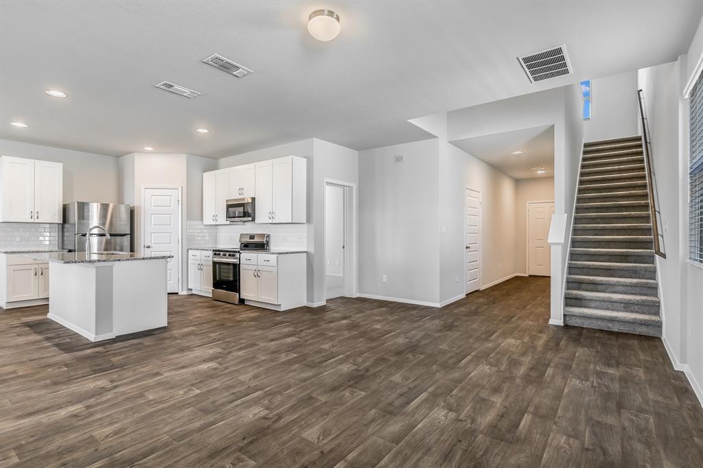 2808 Totley Road Manor, TX 78653 - Photo 4 of 21 Open-concept living space featuring wood-finish flooring, a contemporary kitchen with stainless steel appliances, white cabinetry, a center island with sink, and a staircase with carpeted treads