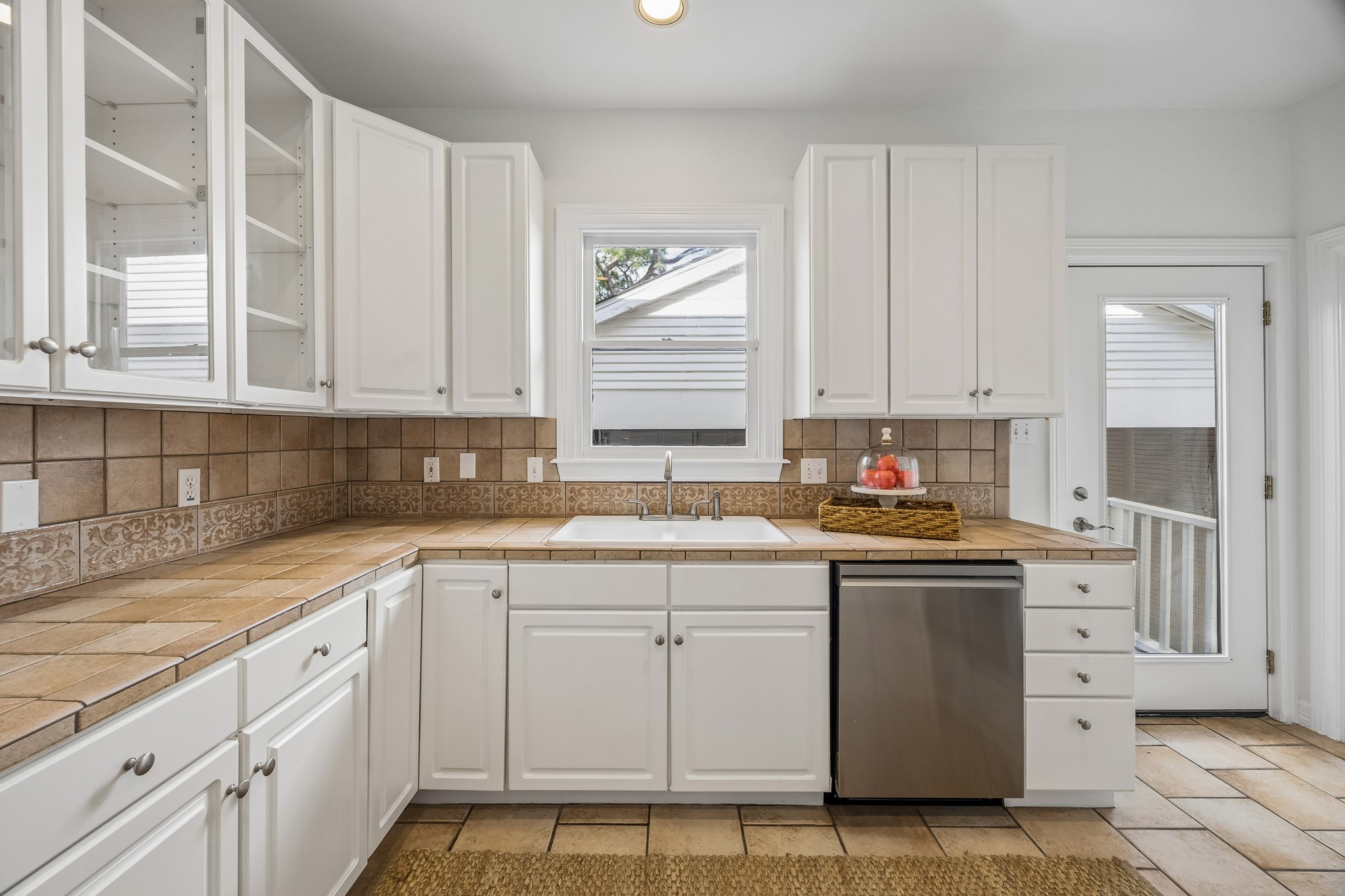 812 West 13th Street Houston, TX 77008 - Photo 13 of 29 This bright kitchen features white cabinetry, a double sink under a window, and a stainless steel dishwasher. The warm tile backsplash and countertops complement the tiled floor, and there's access to an outdoor area through a glass door.