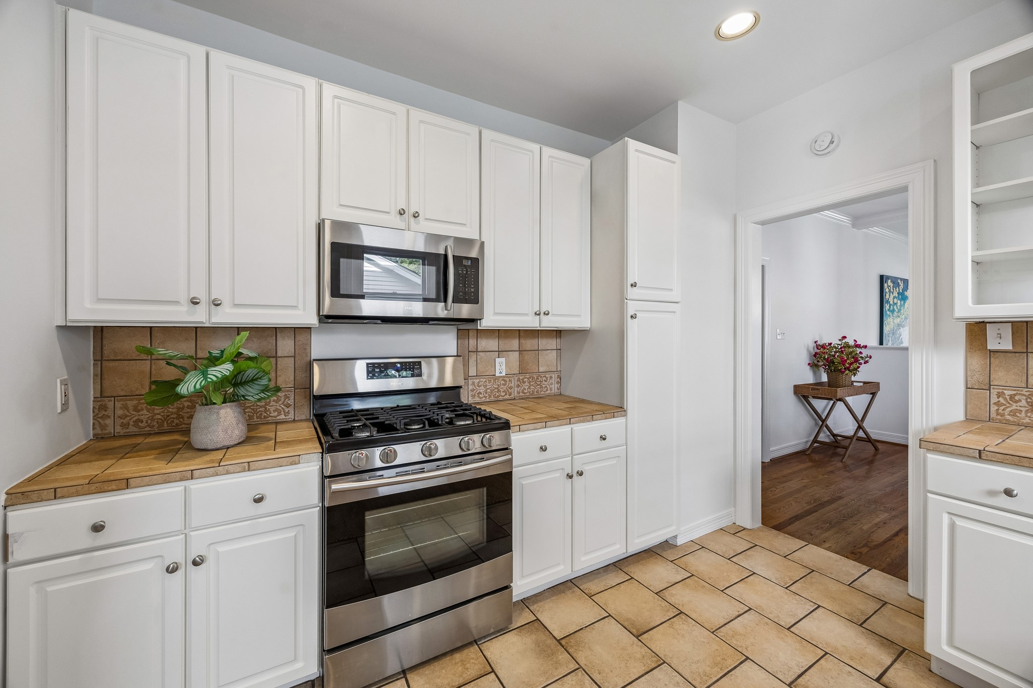 812 West 13th Street Houston, TX 77008 - Photo 15 of 29 This kitchen features white cabinetry, a stainless steel oven and microwave, and tan tile countertops and flooring. It has a clean, bright look with a plant accent and an open doorway leading to a room with hardwood flooring.