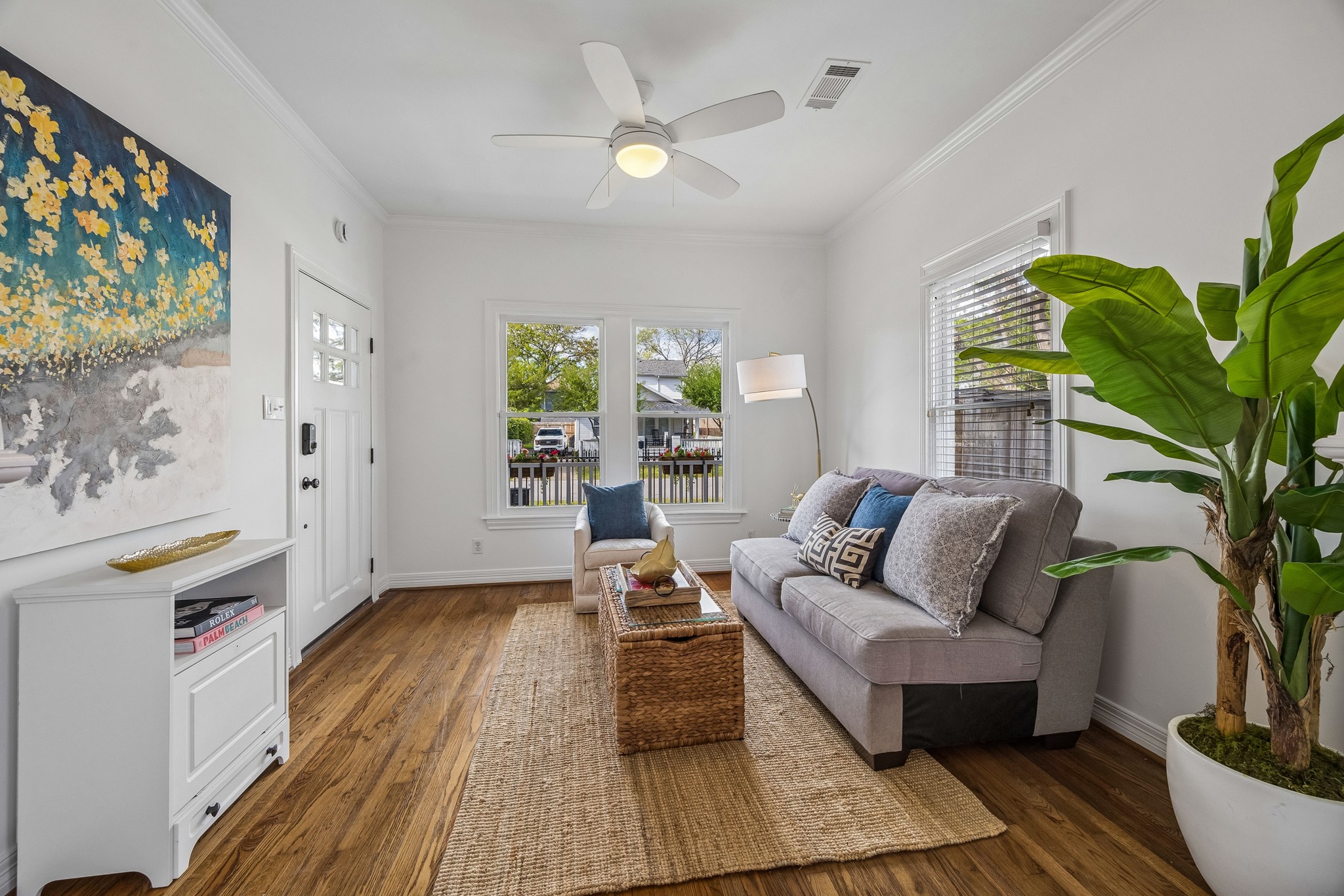 812 West 13th Street Houston, TX 77008 - Photo 2 of 29 This cozy living room features hardwood floors, a large window for natural light, and a modern ceiling fan. It’s tastefully decorated with a comfortable gray sofa, a large plant, and vibrant artwork, creating a welcoming atmosphere.
