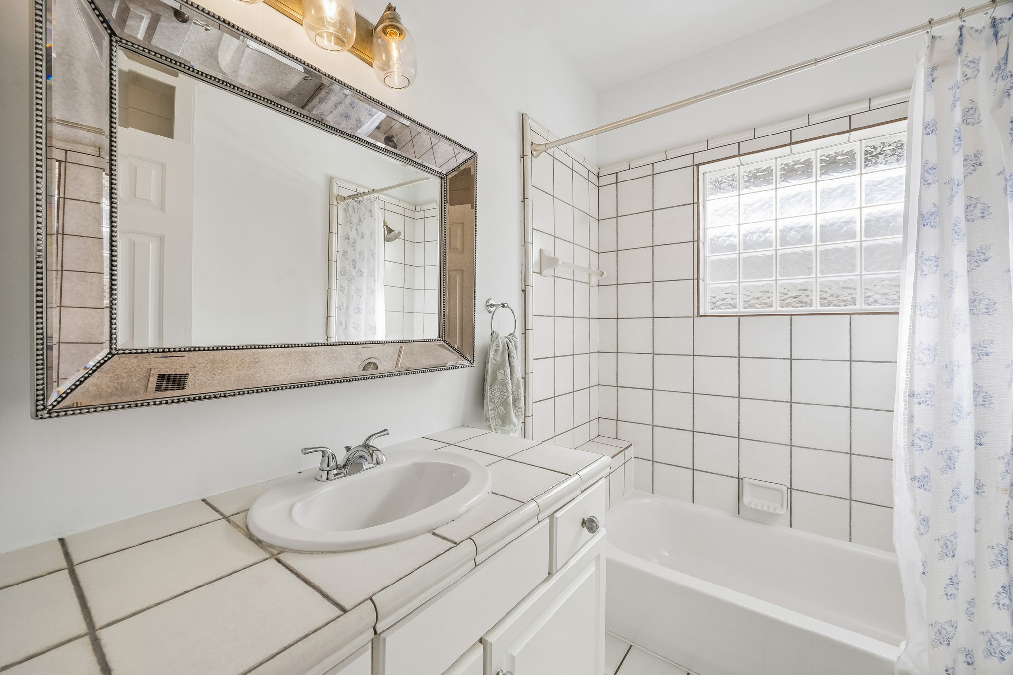 812 West 13th Street Houston, TX 77008 - Photo 21 of 29 This bright bathroom features a large decorative mirror, elegant lighting, tiled countertop, and a bathtub with a classic shower curtain. The glass block window provides natural light, enhancing the spacious feel.