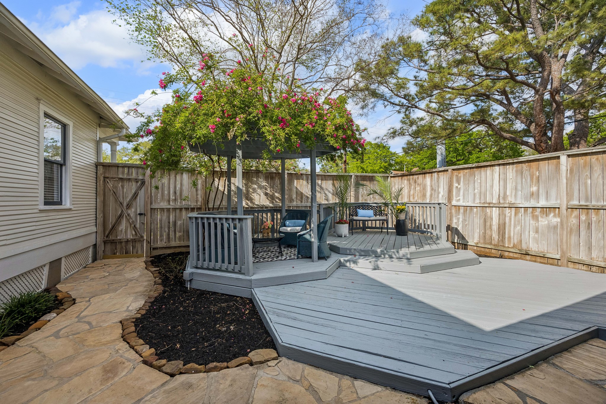 812 West 13th Street Houston, TX 77008 - Photo 22 of 29 This backyard features a charming wooden deck with a pergola adorned with flowering plants, providing a cozy seating area. A stone path leads around the space, enclosed by a wooden fence, offering a private and inviting outdoor retreat.