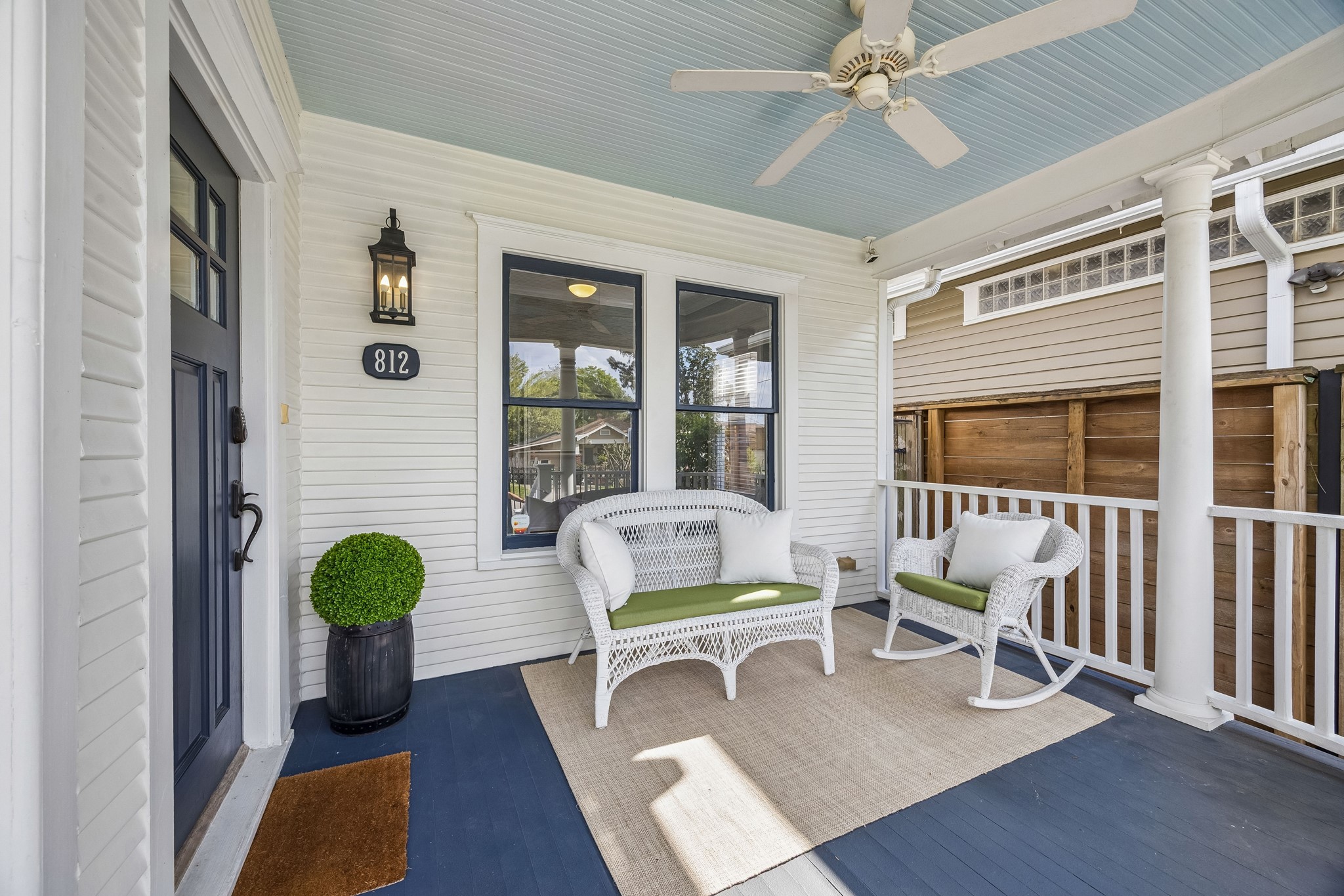 812 West 13th Street Houston, TX 77008 - Photo 9 of 29 This inviting porch features a charming seating area with white wicker furniture, green cushions, and decorative pillows. The blue ceiling and ceiling fan add a touch of elegance, while the potted plant and exterior lighting enhance the welcoming atmosphere. Perfect for relaxing or entertaining.