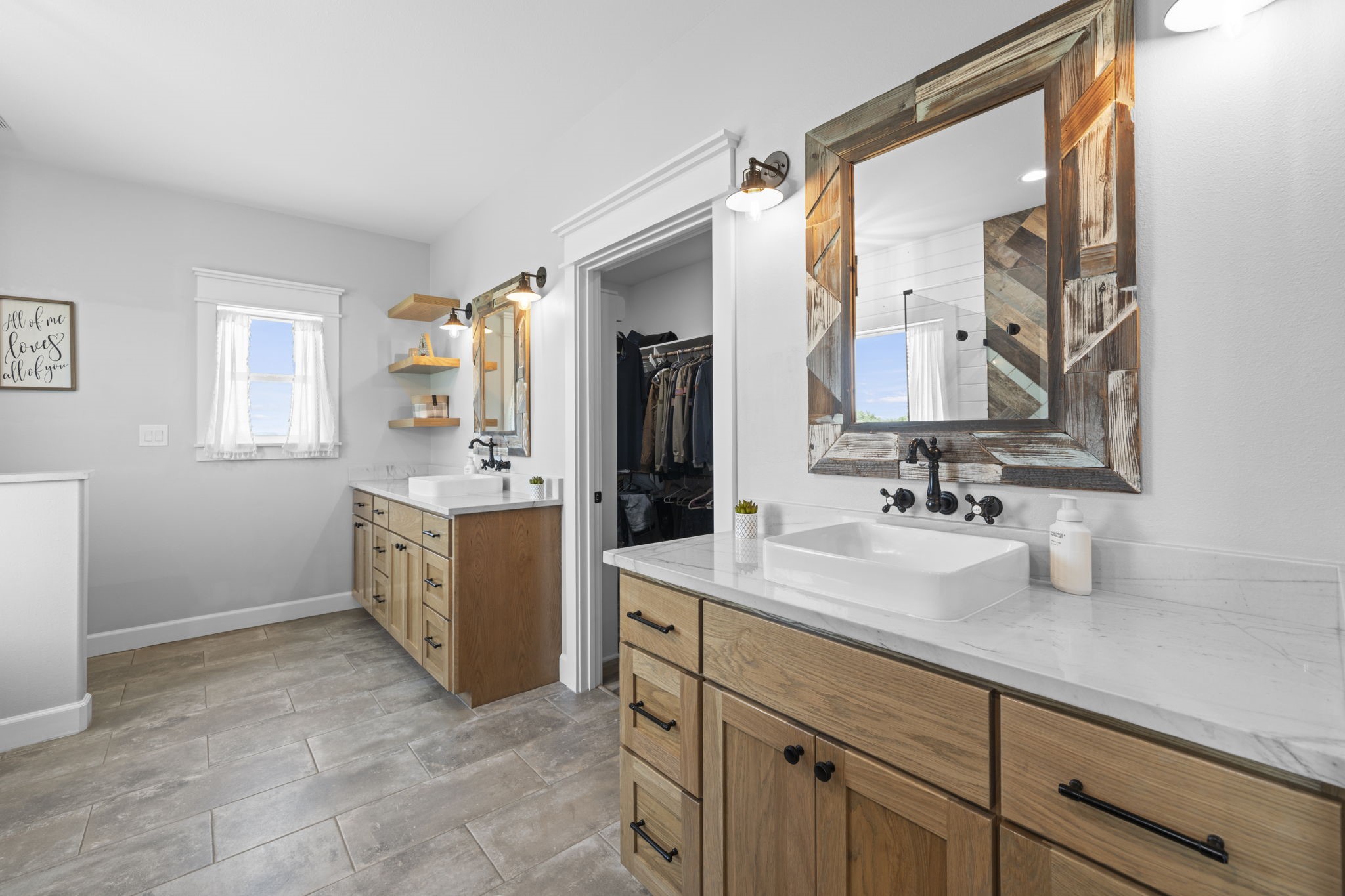 1655 Old Cuero Road Yoakum, TX 77995 - Photo 10 of 24 This luxurious primary bathroom combines modern farmhouse style with exceptional functionality. The spacious dual-sink vanity features custom wood cabinetry, elegant vessel sinks, quartz countertops, and rustic framed mirrors accented by matte black wall-mounted faucets and fixtures. Floating wood shelves provide stylish display and storage space, while abundant natural light creates a bright, spa-like atmosphere.