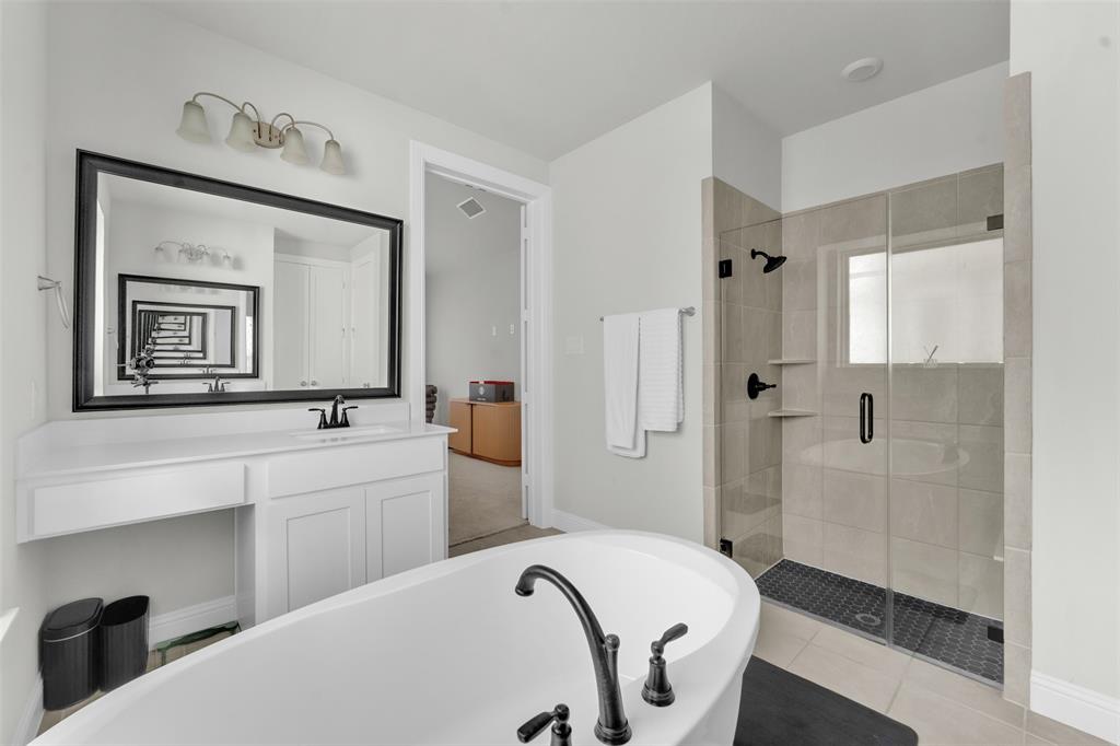 3520 Helios Lane Corinth, TX 76208 - Photo 19 of 37 Contemporary bathroom featuring a freestanding soaking tub with matte black fixtures, a tiled walk-in shower with frameless glass enclosure, a spacious vanity with white cabinetry, a large framed mirror, and light-toned wall tile
