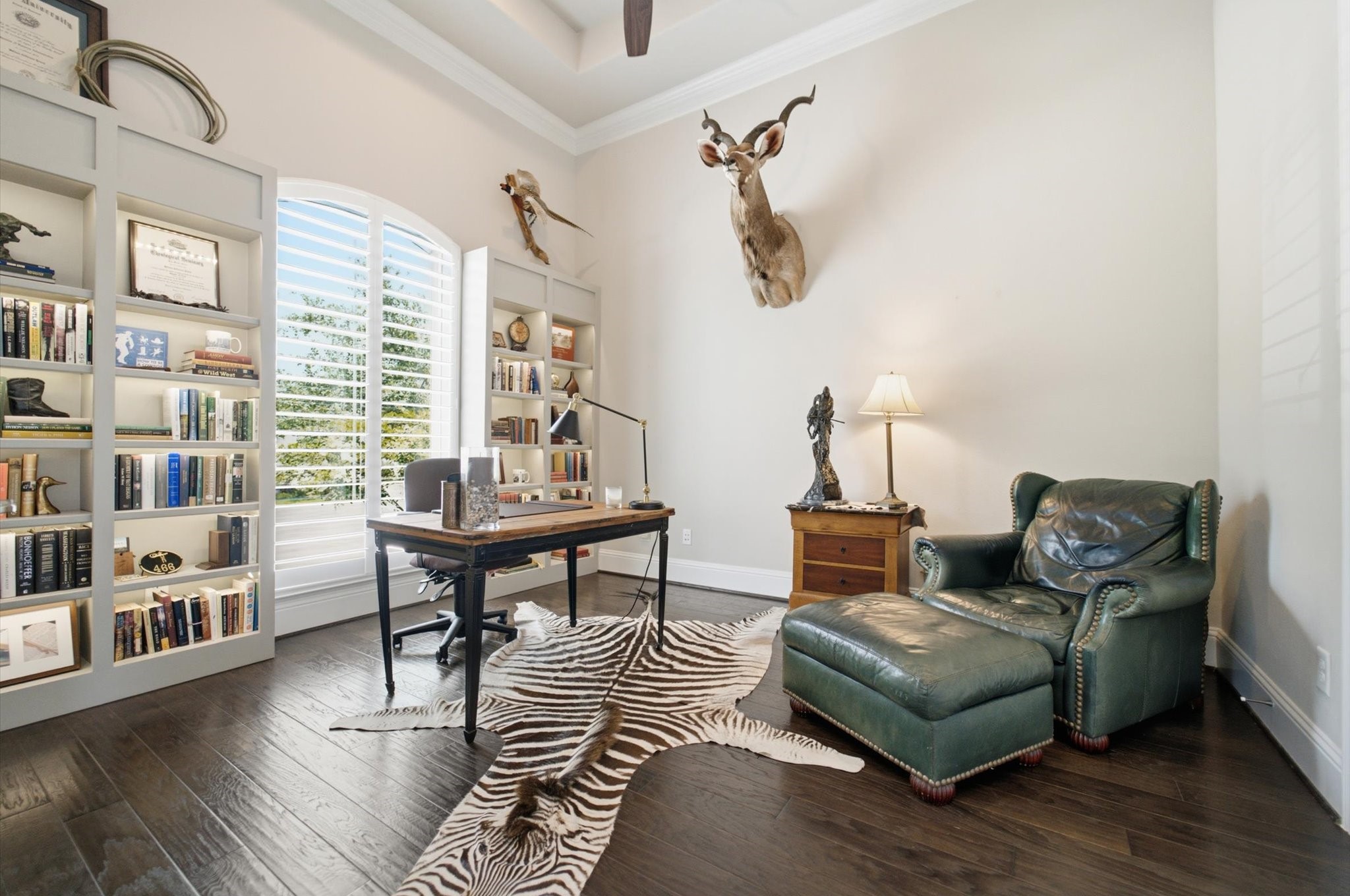 23722 Thortons Park Lane Spring, TX 77389 - Photo 7 of 48 This stylish home office features built-in bookshelves, a large window with shutters, and elegant wood flooring. A classic desk and a comfortable leather armchair with an ottoman provide a cozy workspace. Decor includes a zebra-patterned rug and wall-mounted animal trophies, adding a touch of character.