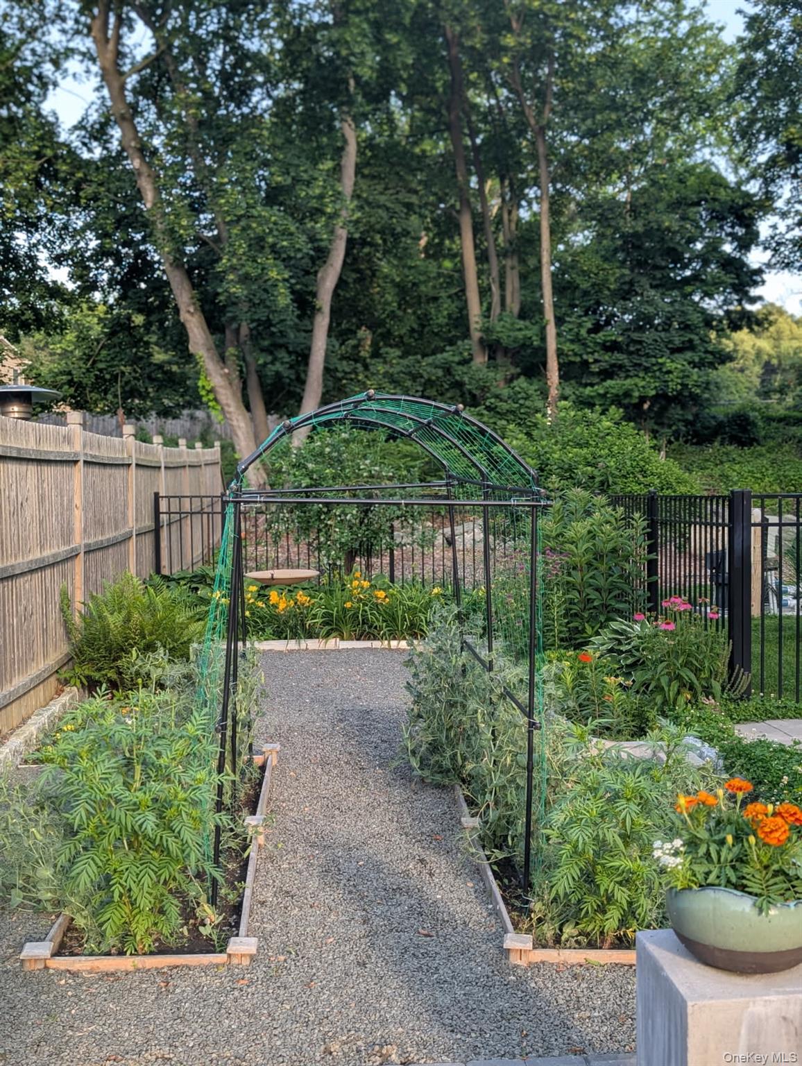 98 Greenbush Road Tappan, NY 10983 - Photo 33 of 33 Landscaped garden pathway featuring a metal archway, gravel ground cover, raised planting beds, wood panel fencing, and mature trees