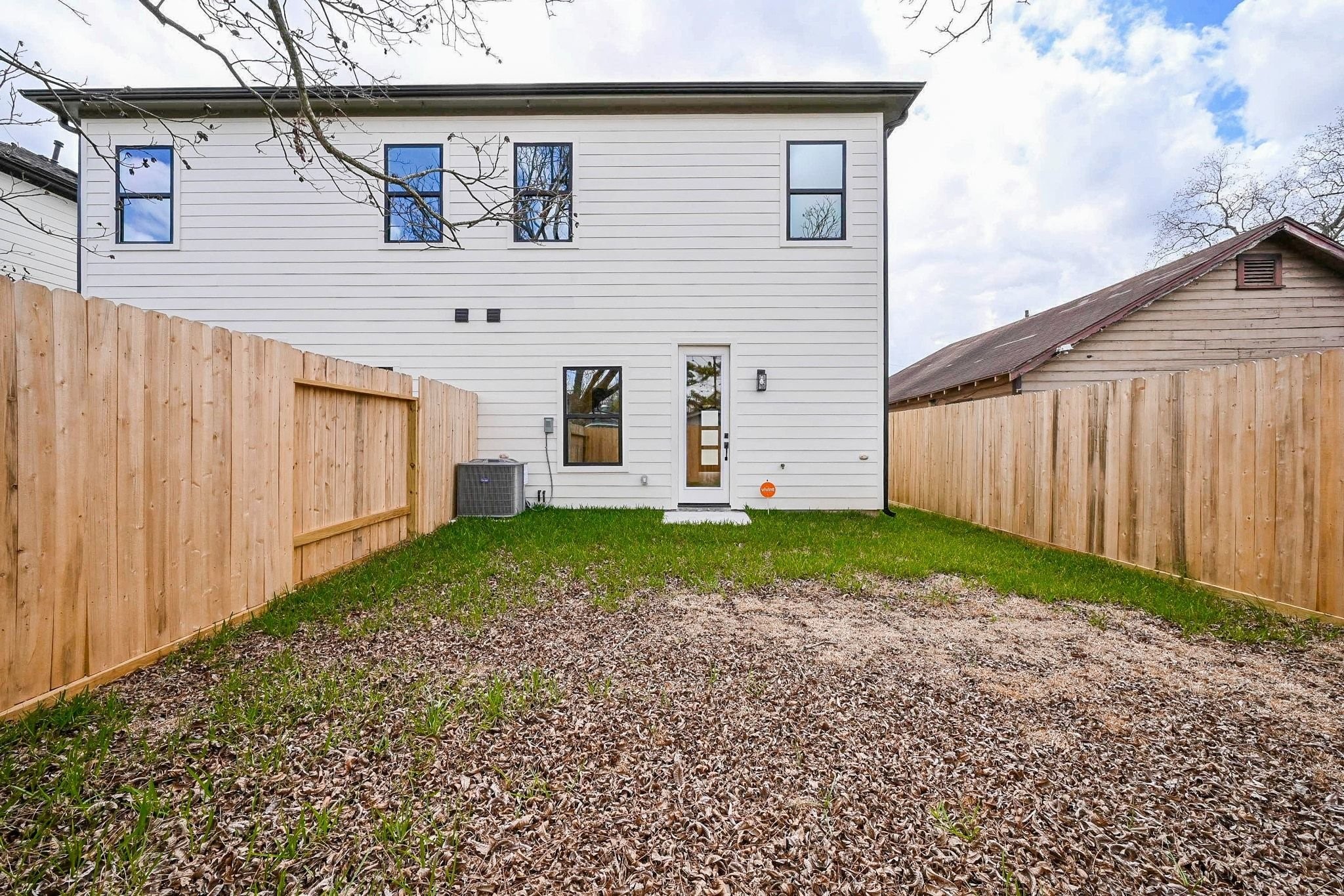 3839 Noah Street, Unit B Houston, TX 77021 - Photo 19 of 19 This photo shows a backyard with a two-story white house featuring a modern door and several windows. The yard is fenced with wood, has a mix of grass and bare patches, and includes an outdoor air conditioning unit.