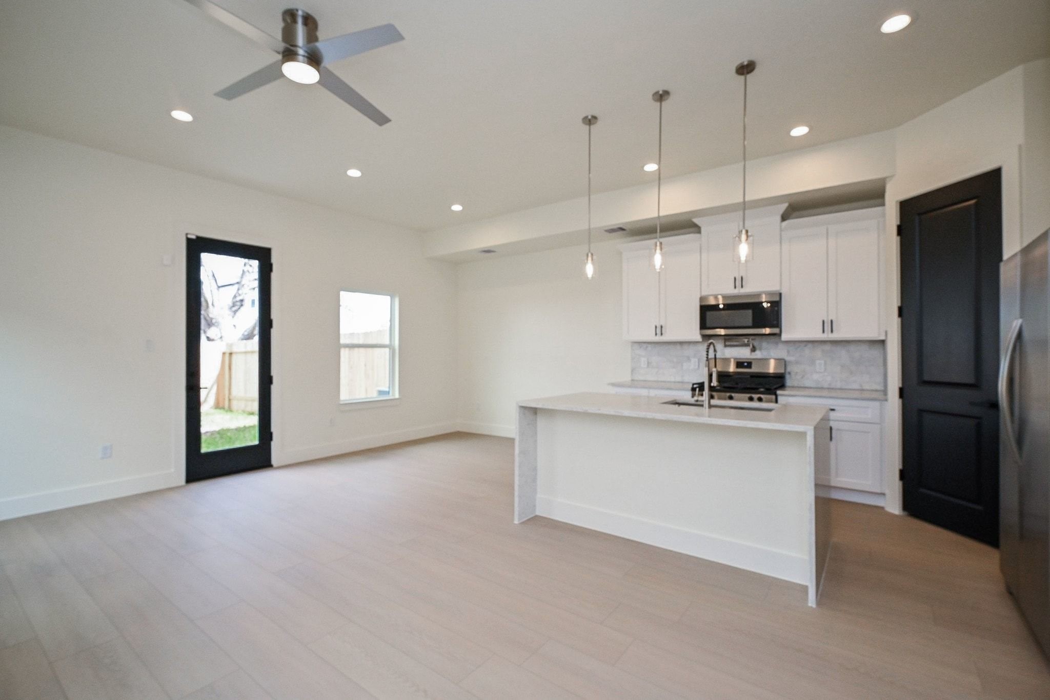 3839 Noah Street, Unit B Houston, TX 77021 - Photo 4 of 19 This photo showcases a modern, open-concept kitchen and living area with light wood flooring. It features a sleek kitchen with white cabinets, a large island, stainless steel appliances, and pendant lighting. The space is bright with natural light from a window and glass door leading to the backyard.
