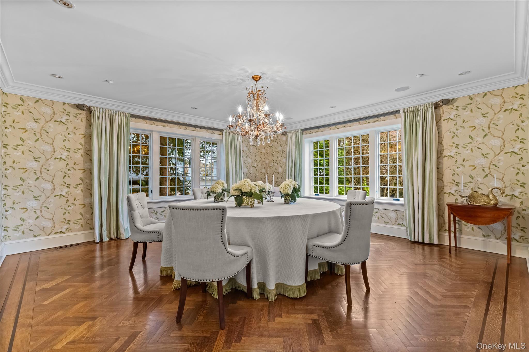 19 Park Drive South Rye, NY 10580 - Photo 17 of 42 Elegant formal dining room featuring a stunning crystal chandelier, patterned wallpaper, and beautiful herringbone hardwood floors. Expansive windows with soft drapery fill the space with natural light, creating an inviting setting for entertaining.