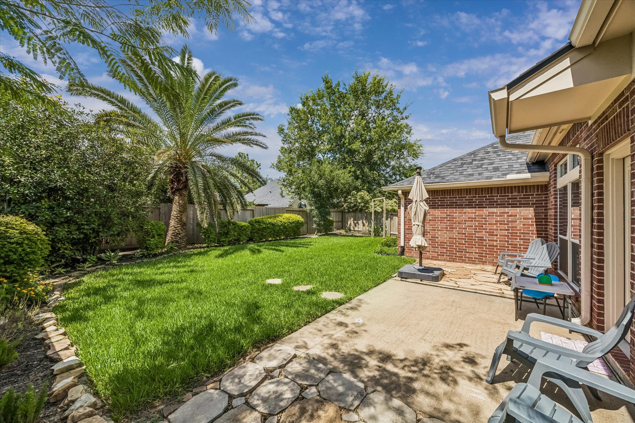 3118 Valley Court Manvel, TX 77578 - Photo 33 of 38 A spacious backyard features a well-maintained lawn with lush greenery and palm trees, bordered by a wooden fence. A brick patio offers seating and an umbrella, ideal for relaxing or entertaining outdoors.