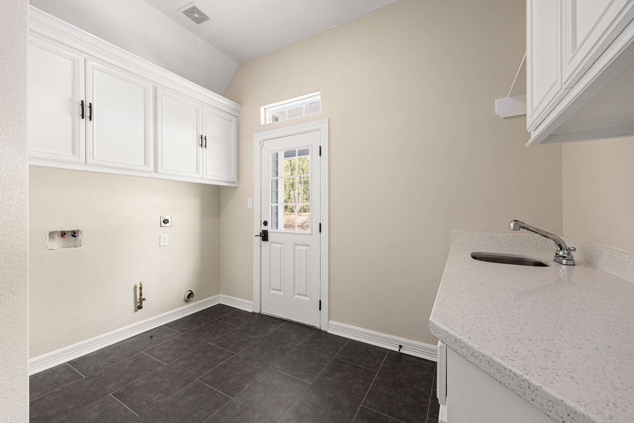 14882 Gingham Road Montgomery, TX 77356 - Photo 21 of 41 This photo shows a bright utility room featuring white cabinets, a modern countertop with a small sink, and dark tiled flooring. The room includes space for a washer and dryer, with a door leading outside.