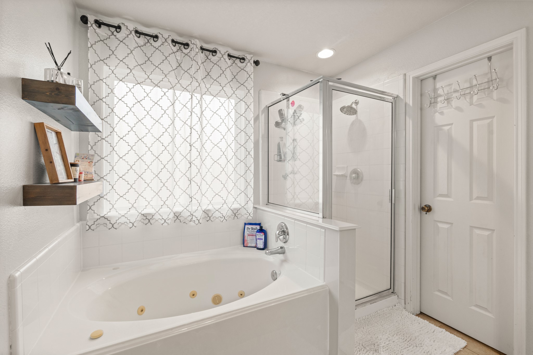 19646 Hardwood Ridge Trail Cypress, TX 77429 - Photo 14 of 22 It features a jetted soaking tub with white tile surround, positioned beneath a window that allows natural light to fill the space while maintaining privacy through patterned curtains. Adjacent to the tub is a separate glass-enclosed shower, offering a practical and modern layout.