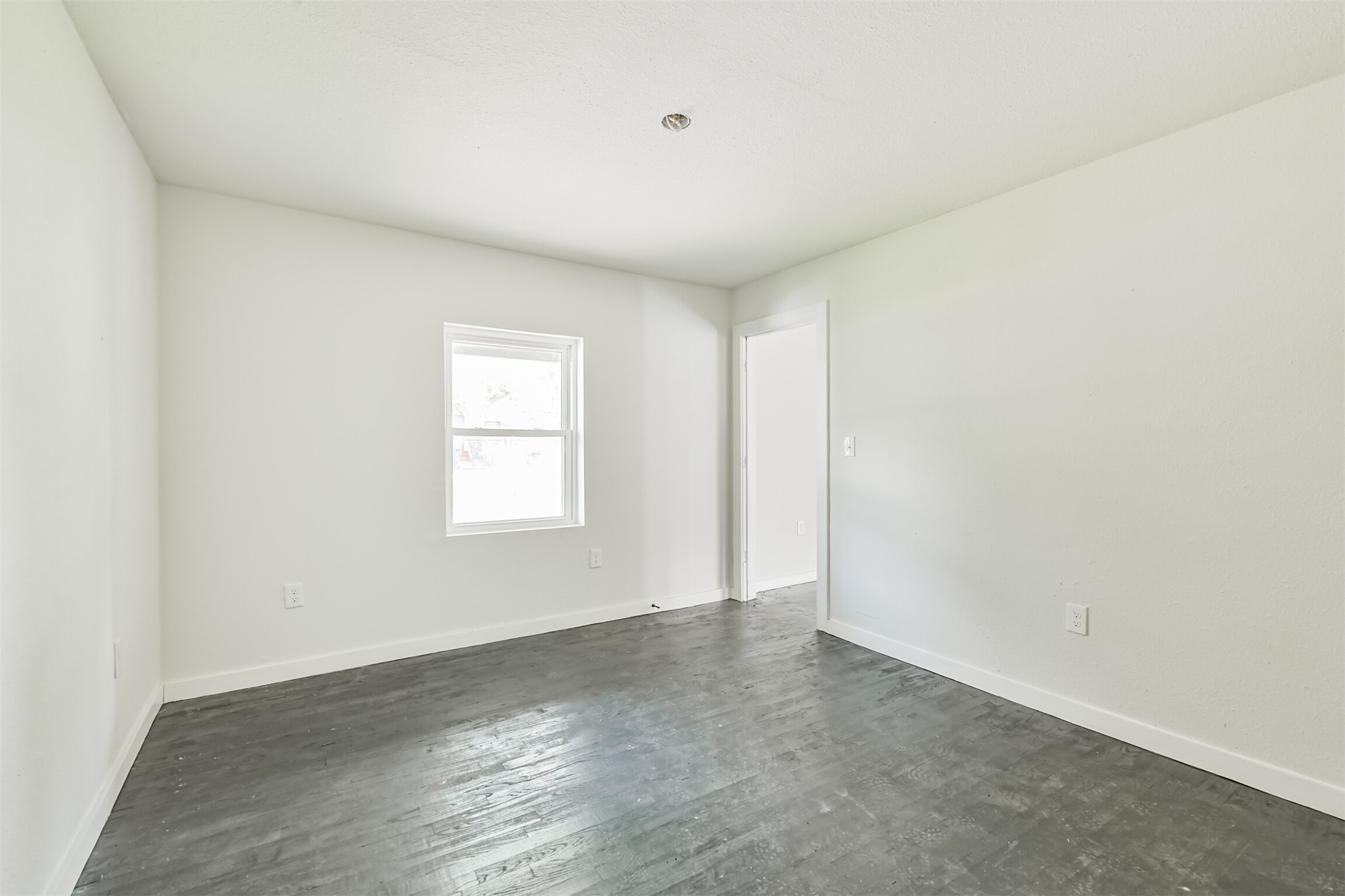 209 Leago Street Houston, TX 77022 - Photo 12 of 24 This photo shows a bright, empty room with light gray walls and dark wood flooring. It features a window and a doorway, offering a clean and modern space.