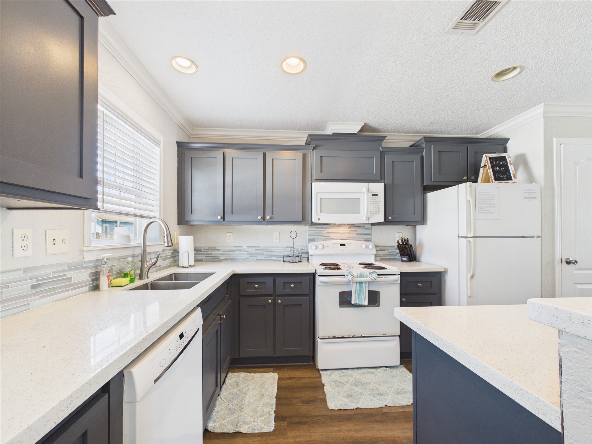 905 Meynig Drive Crystal Beach, TX 77650 - Photo 17 of 50 The kitchen features modern dark cabinetry with white countertops and appliances, including a dishwasher, microwave, and refrigerator. It has a clean, bright design with a window for natural light, recessed lighting, and a tiled backsplash.