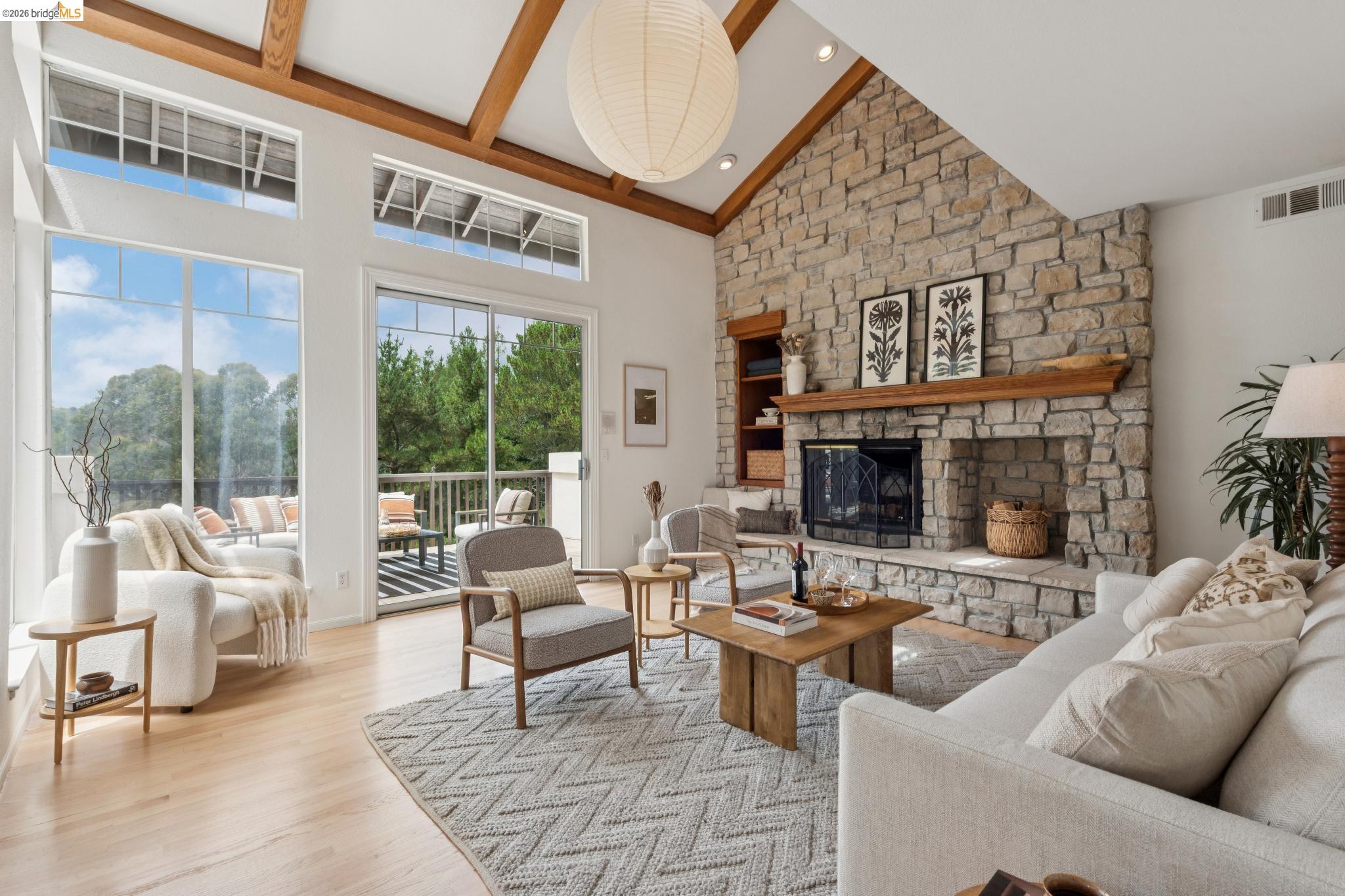1325 Drury Road Berkeley, CA 94705 - Photo 12 of 71 Living area featuring a floor-to-ceiling stone fireplace with a wood mantle and hearth, vaulted ceiling with exposed wood beams, large windows, sliding glass door to an outdoor deck, and wood-finish flooring