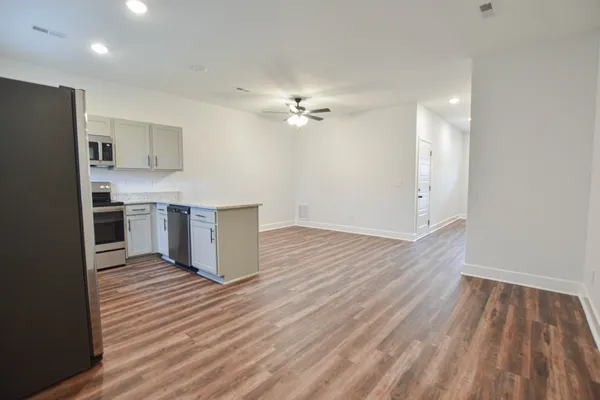 an empty room with wooden floor a ceiling fan a kitchen view and a refrigerator