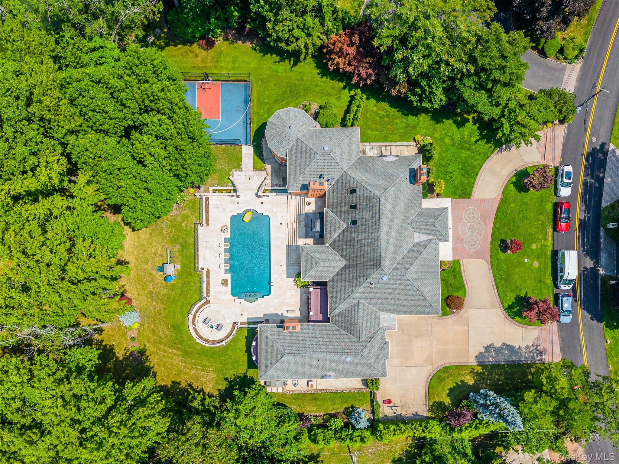 25 Lake Road Lake Success, NY 11020 - Photo 11 of 50 Expansive property featuring a large residence with a multi-level roofline, a winding driveway with paver accents, an integrated swimming pool with a surrounding patio, a dedicated sports court, and mature landscaping