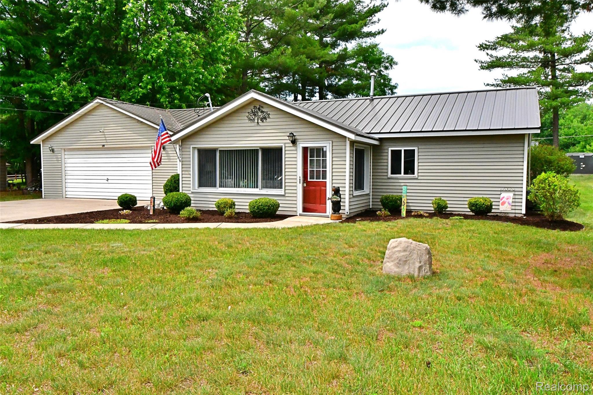 9611 Monroe Road St. Helen, MI 48656 - Photo 1 of 24 Ranch-style exterior featuring light-colored siding, a gray metal roof, an attached two-car garage with a white door, a red front entry door, and a landscaped front yard with decorative shrubs