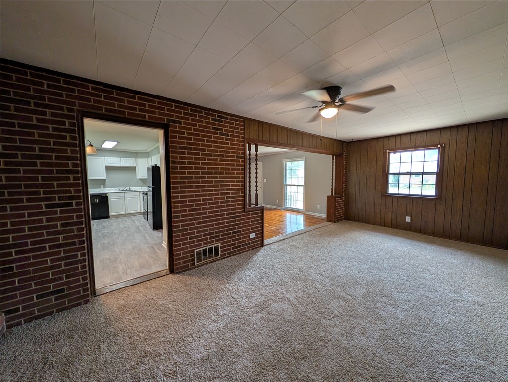 203 Glendale Road Anderson, SC 29624 - Photo 7 of 20 A well-lit living area featuring rich wood paneling, a ceiling fan, and carpeted flooring, with a glimpse into the kitchen and an adjacent room.