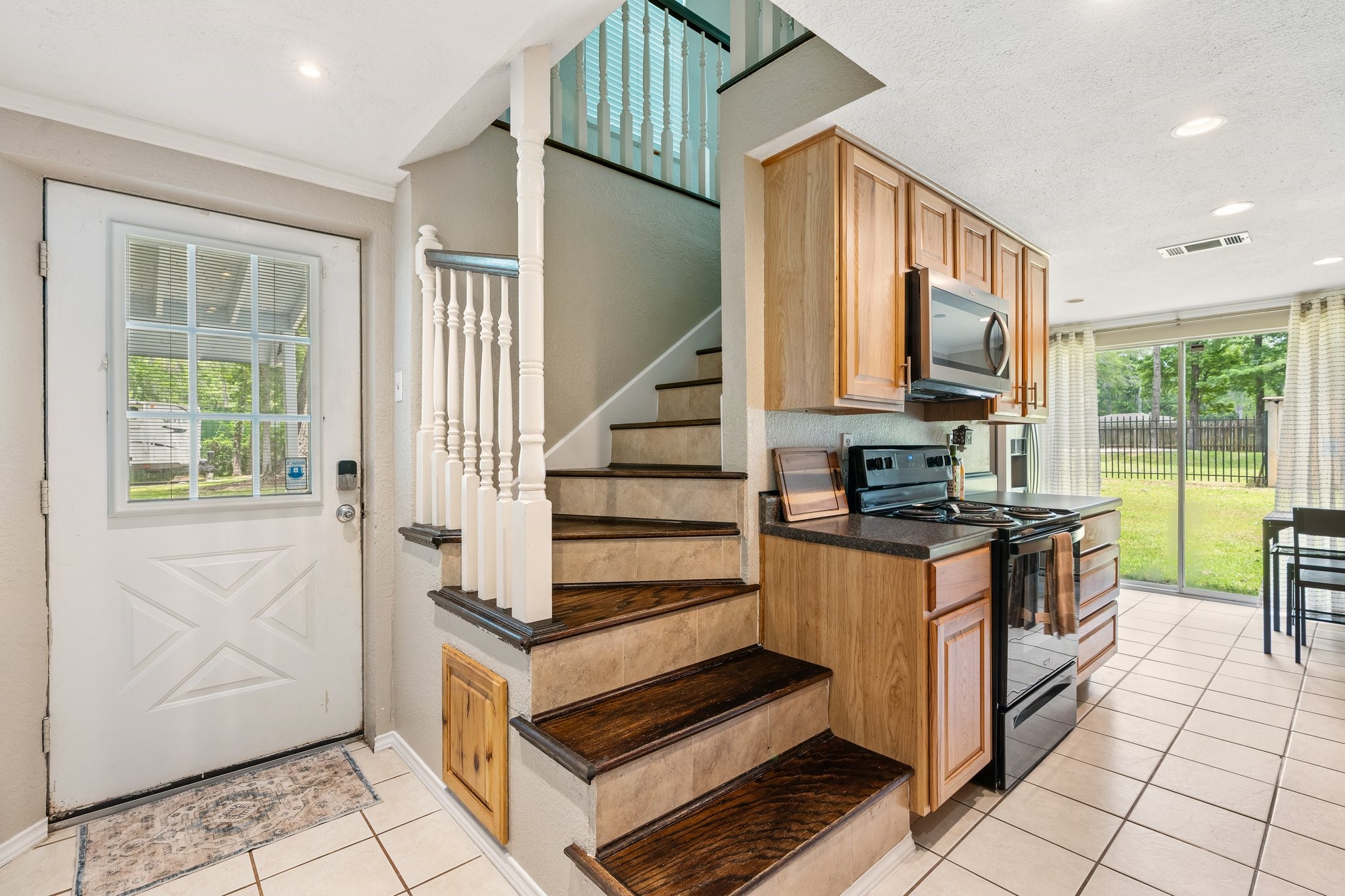 14758 Kirbee Drive Conroe, TX 77302 - Photo 19 of 35 Bright and welcoming kitchen area with wooden cabinets and modern appliances. The space features a staircase with elegant railing, leading to the upper floor. Large sliding glass doors open to a fenced backyard, offering plenty of natural light and easy access to outdoor space. The tiled floor adds a clean and stylish touch.