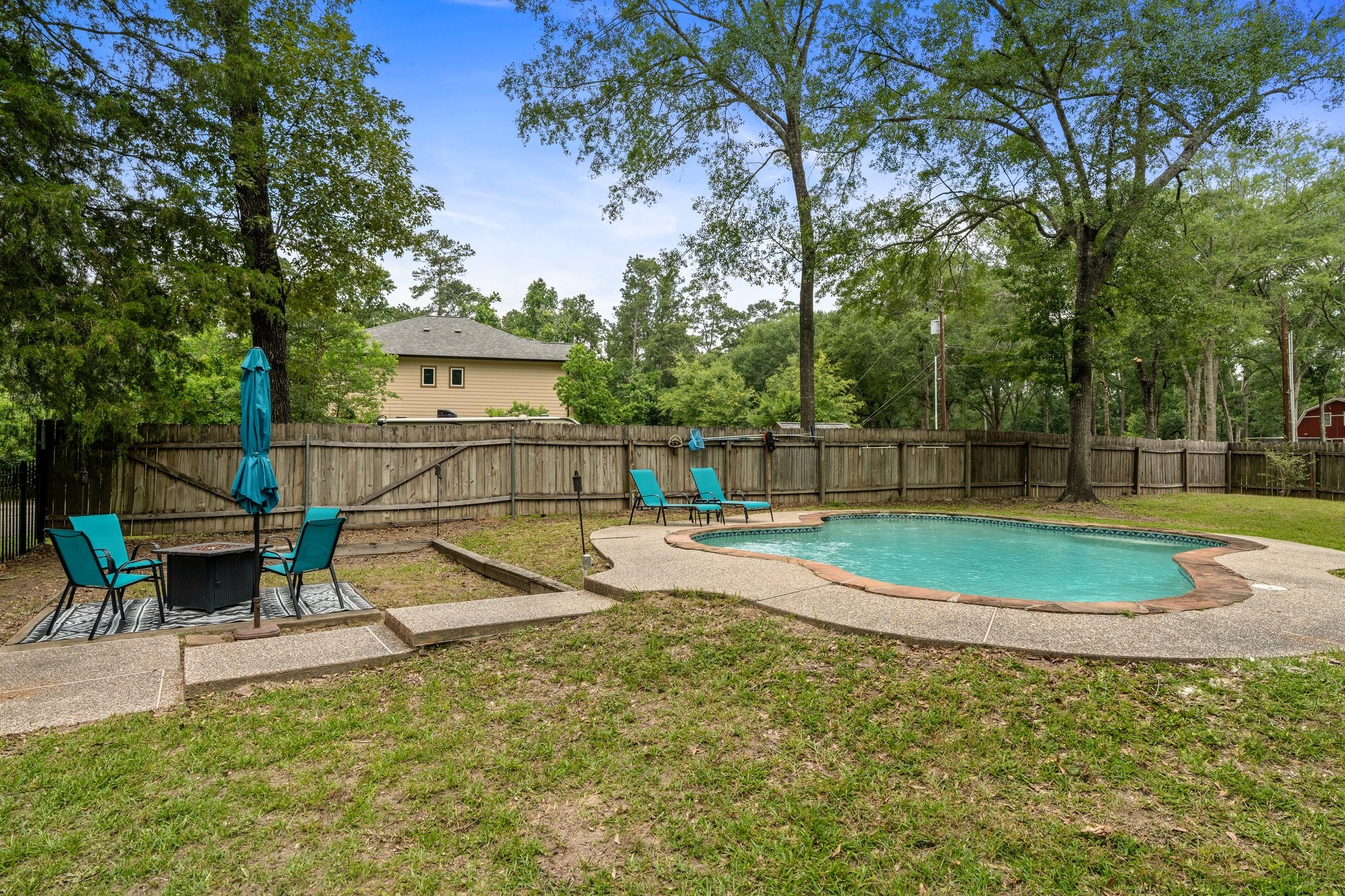 14758 Kirbee Drive Conroe, TX 77302 - Photo 2 of 35 Spacious yard features a private pool with surrounding patio space, set in a fenced area. There's a cozy seating area with a table and umbrella, perfect for outdoor relaxation. Lush greenery provides a serene backdrop.