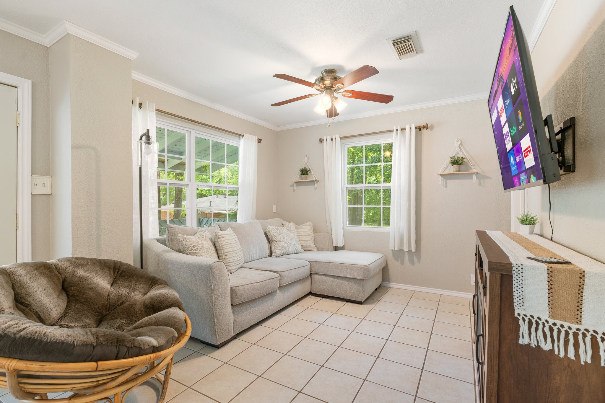 14758 Kirbee Drive Conroe, TX 77302 - Photo 23 of 35 This cozy living room features tiled flooring, a comfortable sectional sofa, and a wall-mounted TV. Natural light streams in through large windows with white curtains. A ceiling fan provides airflow, and the space is tastefully decorated with small shelves and a plush chair.