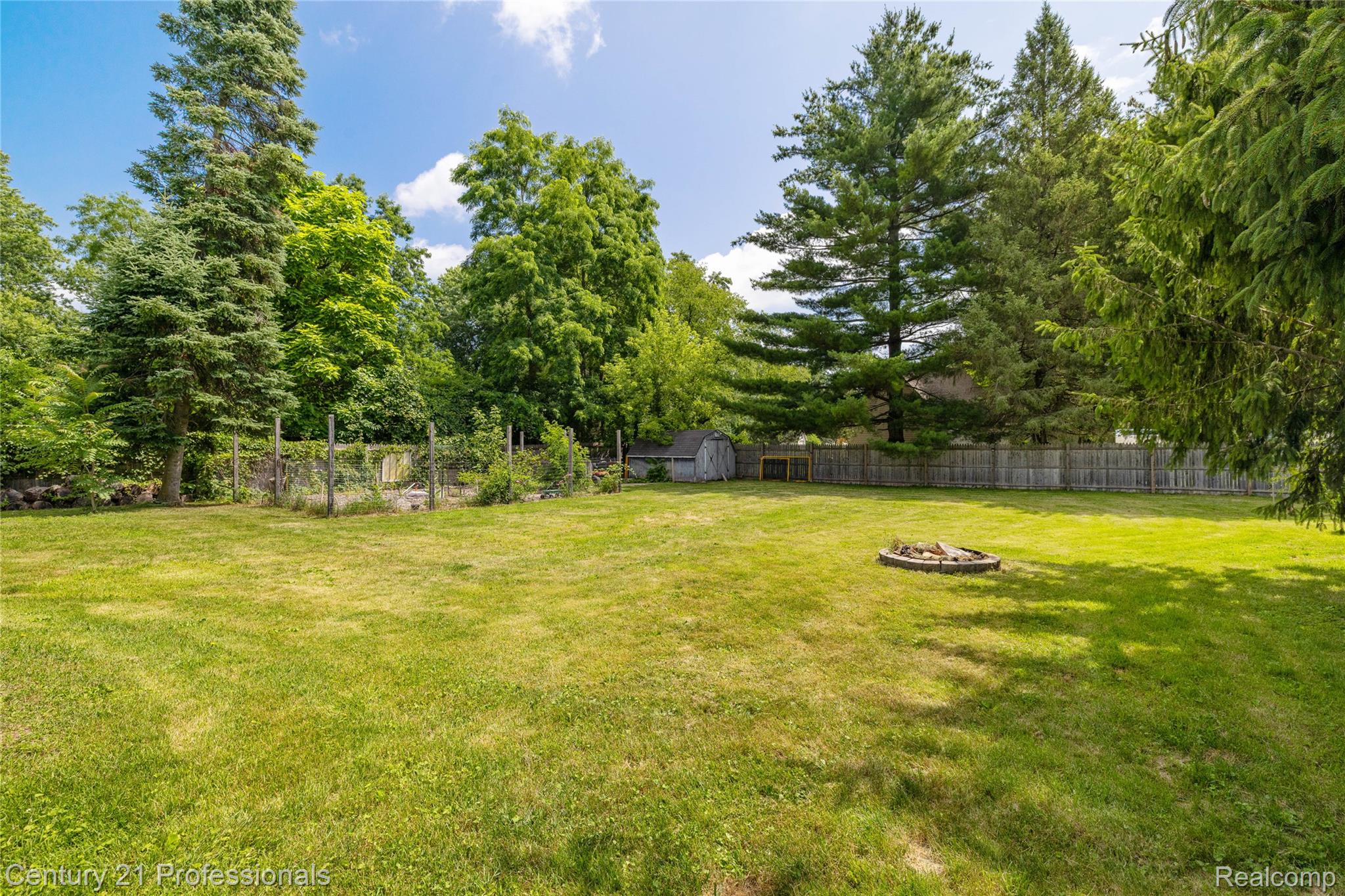 15037 Hogan Road Linden, MI 48451 - Photo 32 of 38 Expansive green lawn featuring a built-in fire pit, a garden area with trellises, a wood privacy fence, a storage shed, and mature trees