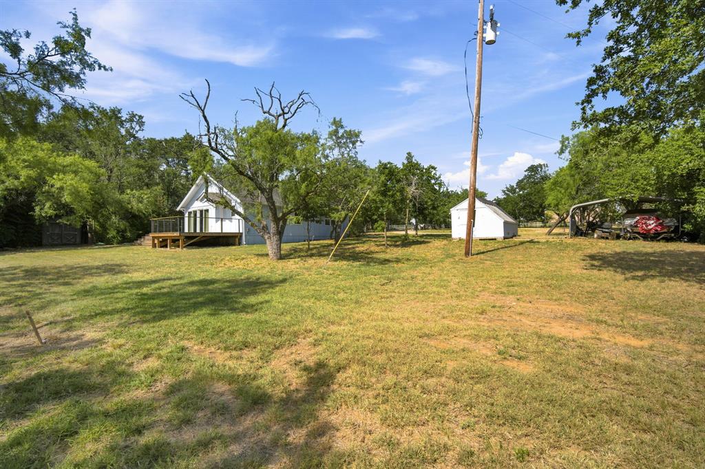 3325 Dove Creek Road Cleburne, TX 76031 - Photo 20 of 40 Expansive grassy outdoor space featuring mature trees, a white residential structure with a raised wooden deck, a detached white shed, and a covered outdoor storage area