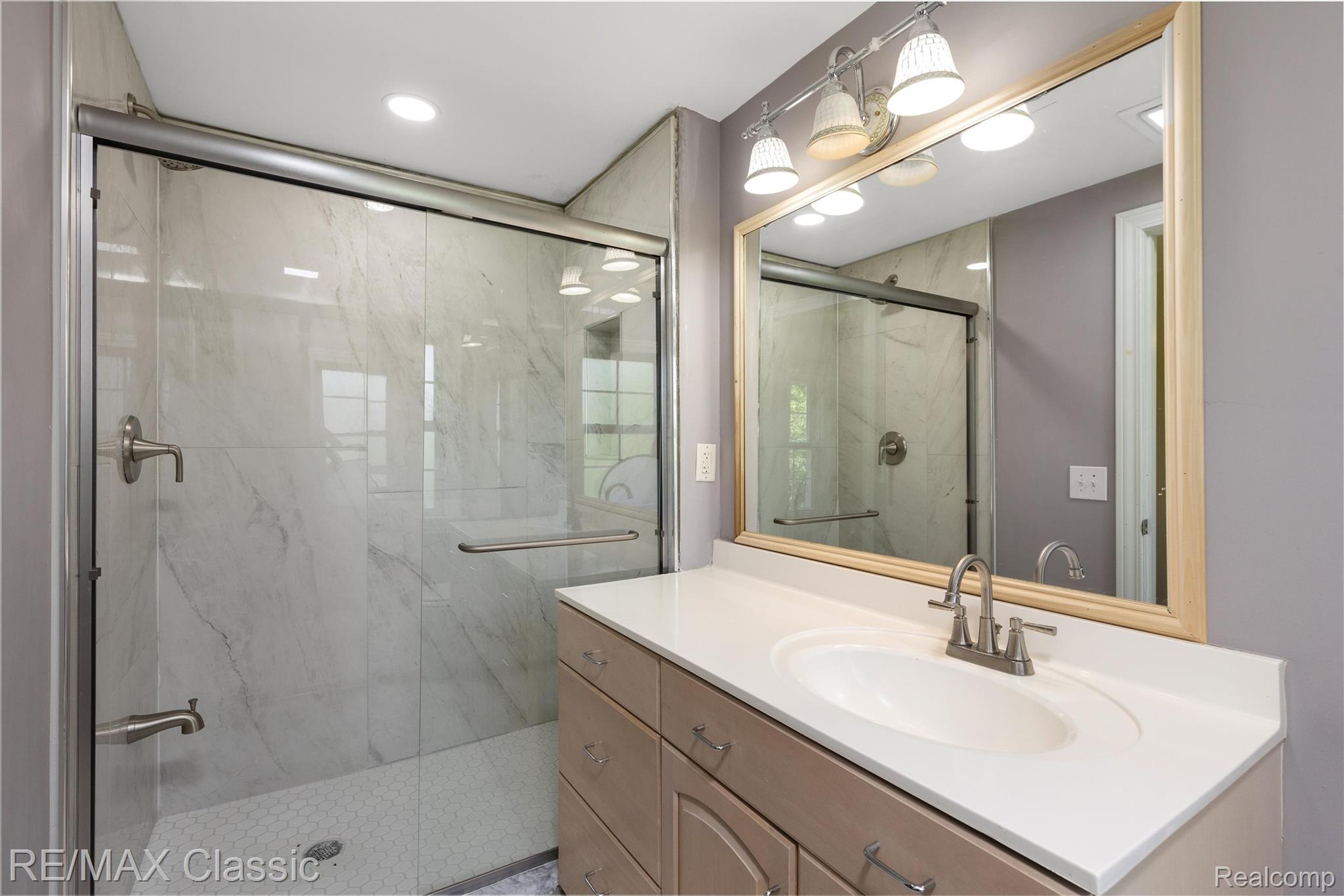 4268 Covered Bridge Road Bloomfield Hills, MI 48302 - Photo 15 of 35 Bathroom featuring a glass-enclosed shower with white marble-look tile, a vanity with a white countertop, an oval undermount sink, and a large framed mirror with overhead lighting