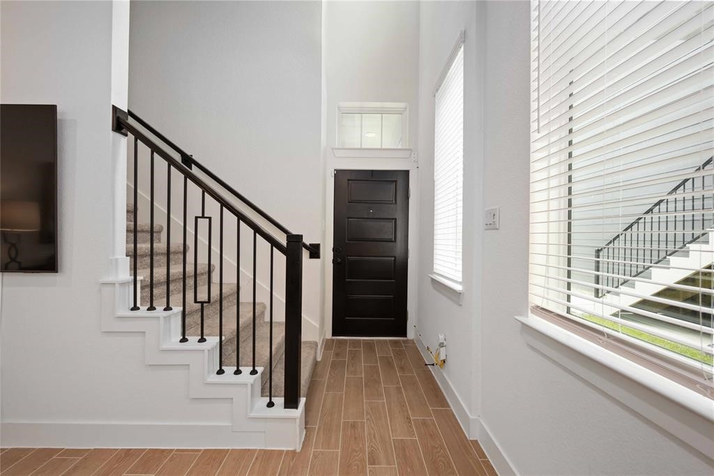 10014 Nectar Path Iowa Colony, TX 77583 - Photo 4 of 26 Bright entryway with high ceilings, featuring a modern staircase with black railings. The space includes large windows with blinds, and a sleek black front door. Wood-look tile flooring adds warmth and style.
