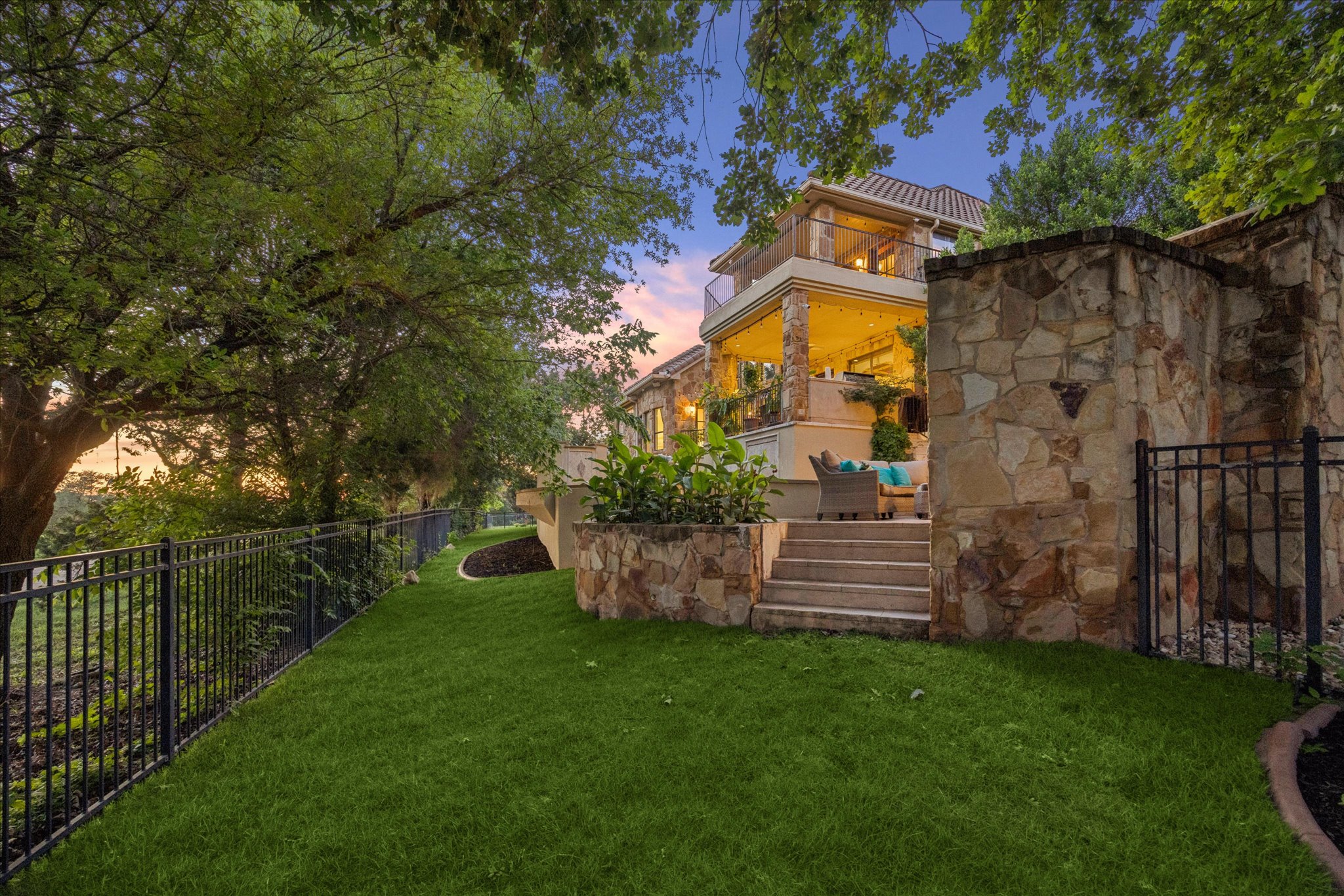 11805 Ranchview Court Austin, TX 78732 - Photo 32 of 36 Multi-level exterior featuring natural stone cladding, a tiered patio with string lighting, a second-story balcony with glass railings, and a fenced yard with mature trees