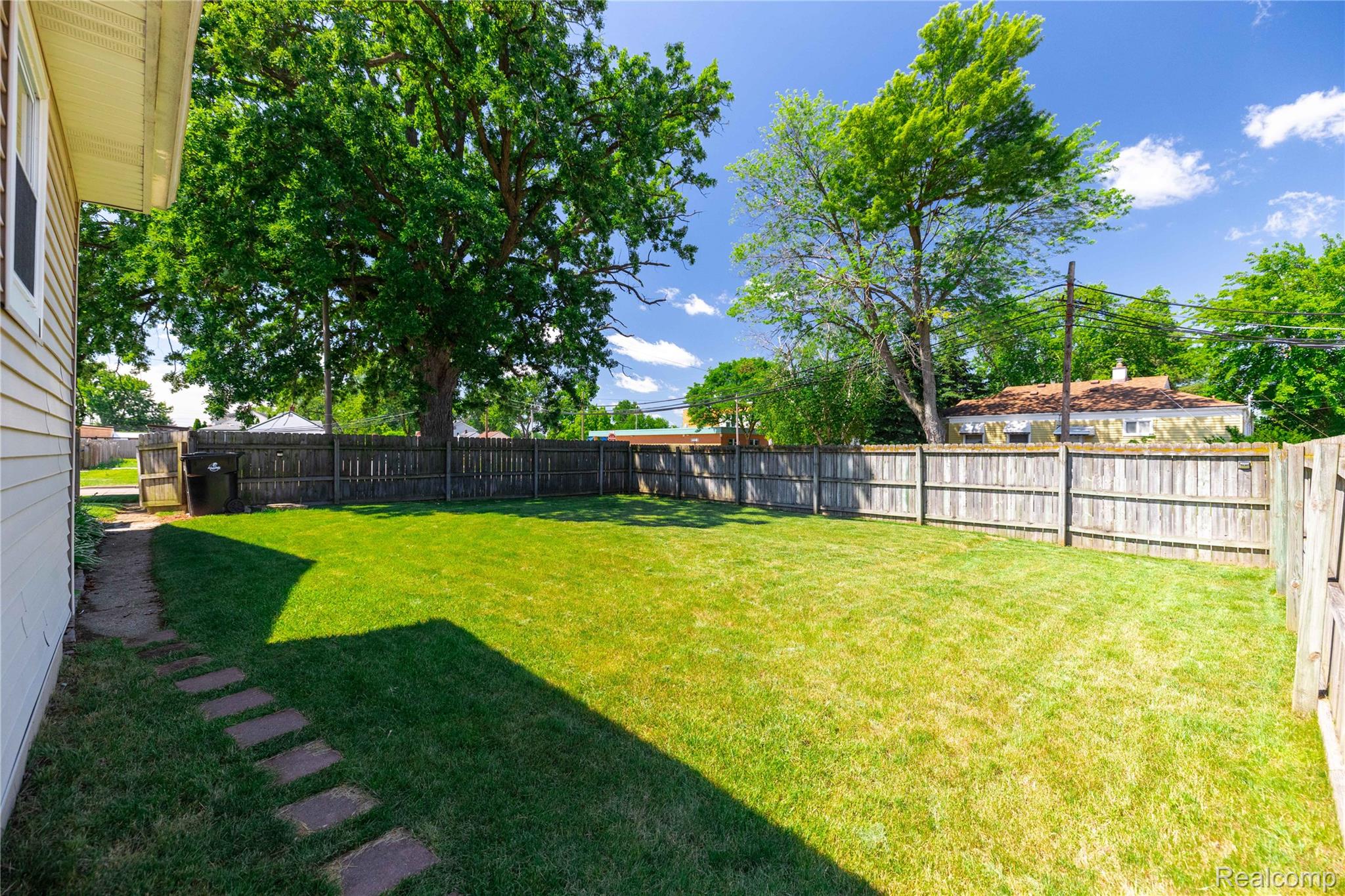 826 Goddard Road Wyandotte, MI 48192 - Photo 29 of 39 Expansive backyard featuring a lush, manicured lawn with mature shade trees, wooden privacy fencing, and a stone-paved pathway along the home's exterior