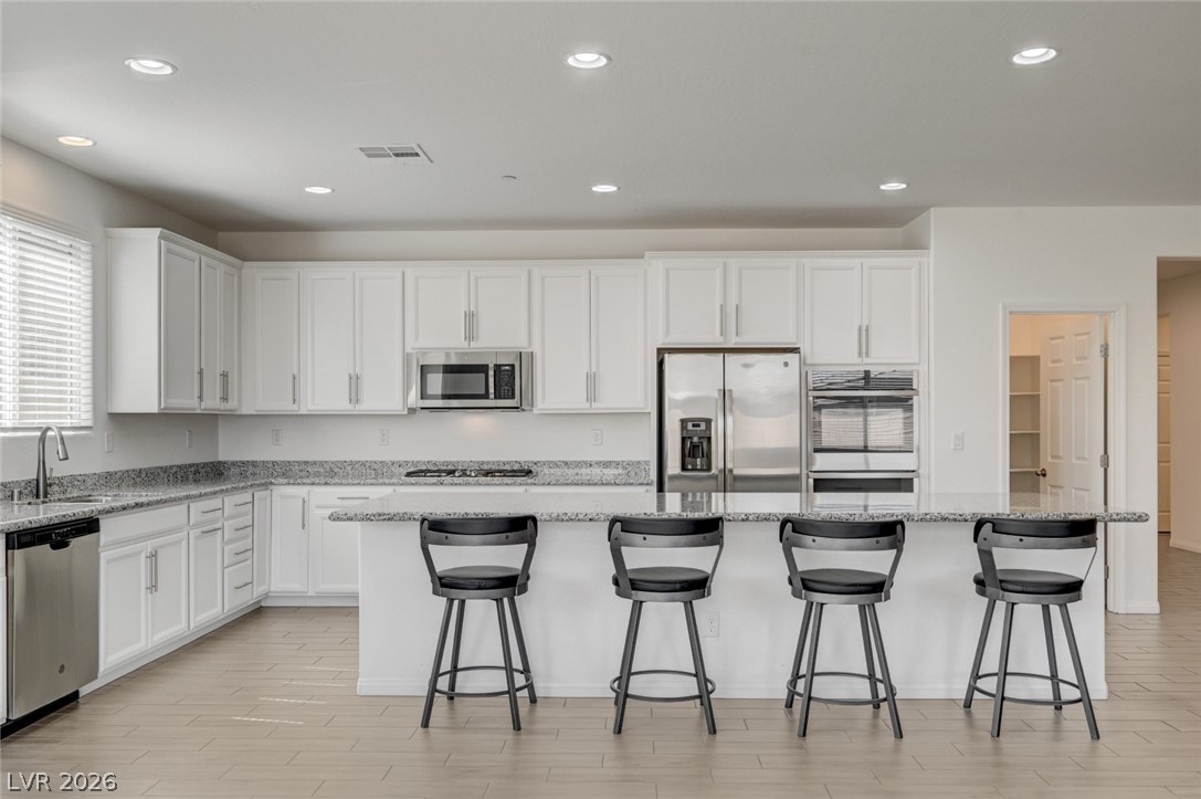 756 Cadence View Way Henderson, NV 89011 - Photo 6 of 45 Kitchen featuring white cabinetry, stainless steel appliances, a kitchen bar, light stone countertops, and wood finish floors
