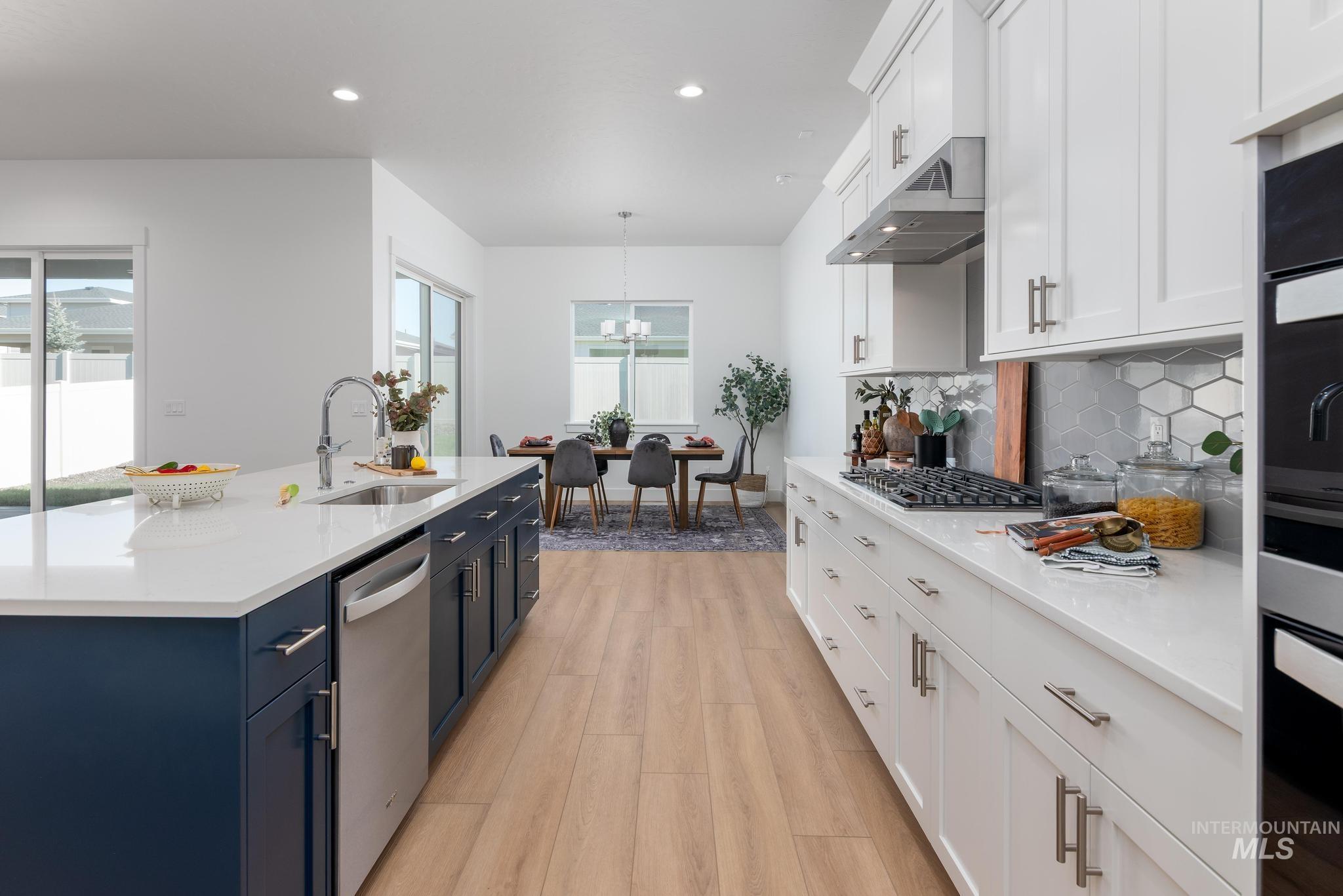9074 West Moonglade Street Star, ID 83669 - Photo 11 of 40 Modern kitchen featuring a large central island with a stainless steel sink and dishwasher, white quartz countertops, white shaker cabinetry, a gas range with stainless steel hood, and wood-finish flooring