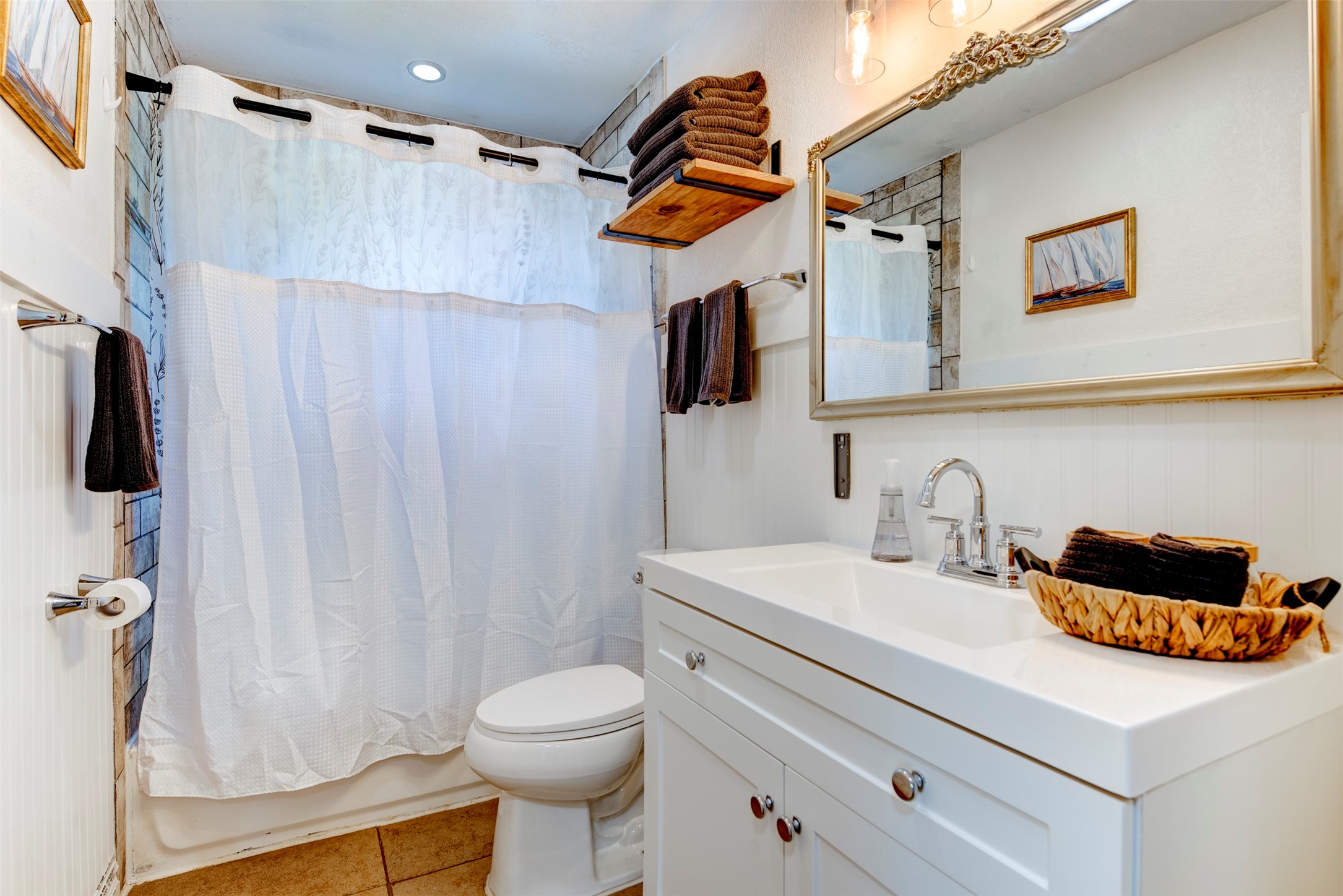 1121 Panama Street Houston, TX 77009 - Photo 20 of 35 Full bathroom featuring white shaker vanity with chrome two-handle faucet, ornate silver-framed mirror, Edison bulb pendant lighting, open natural wood floating shelf, beadboard wainscoting, recessed ceiling light, and tiled shower surround.