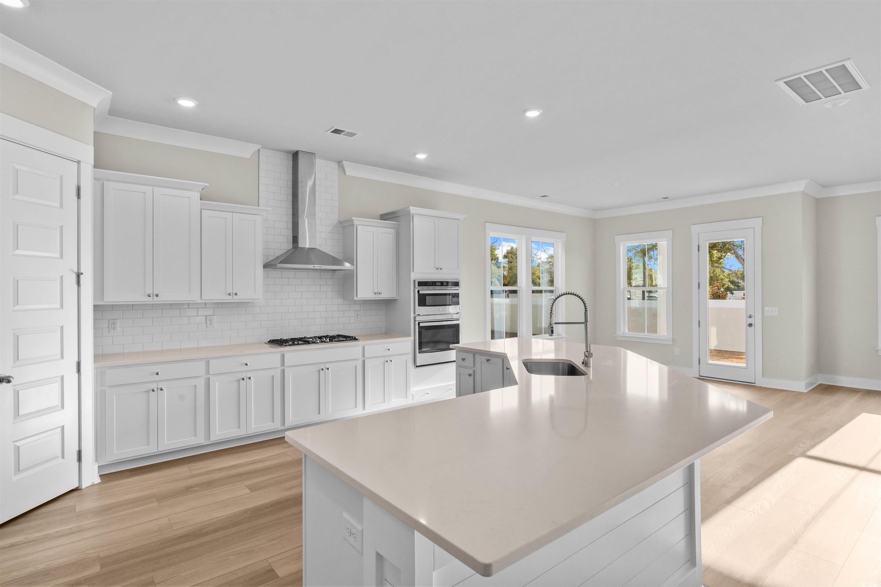 205 Atlantic Breeze Street Myrtle Beach, SC 29572 - Photo 7 of 28 Kitchen with white cabinets, an island with sink, crown molding, light wood-style flooring, and wall chimney exhaust hood