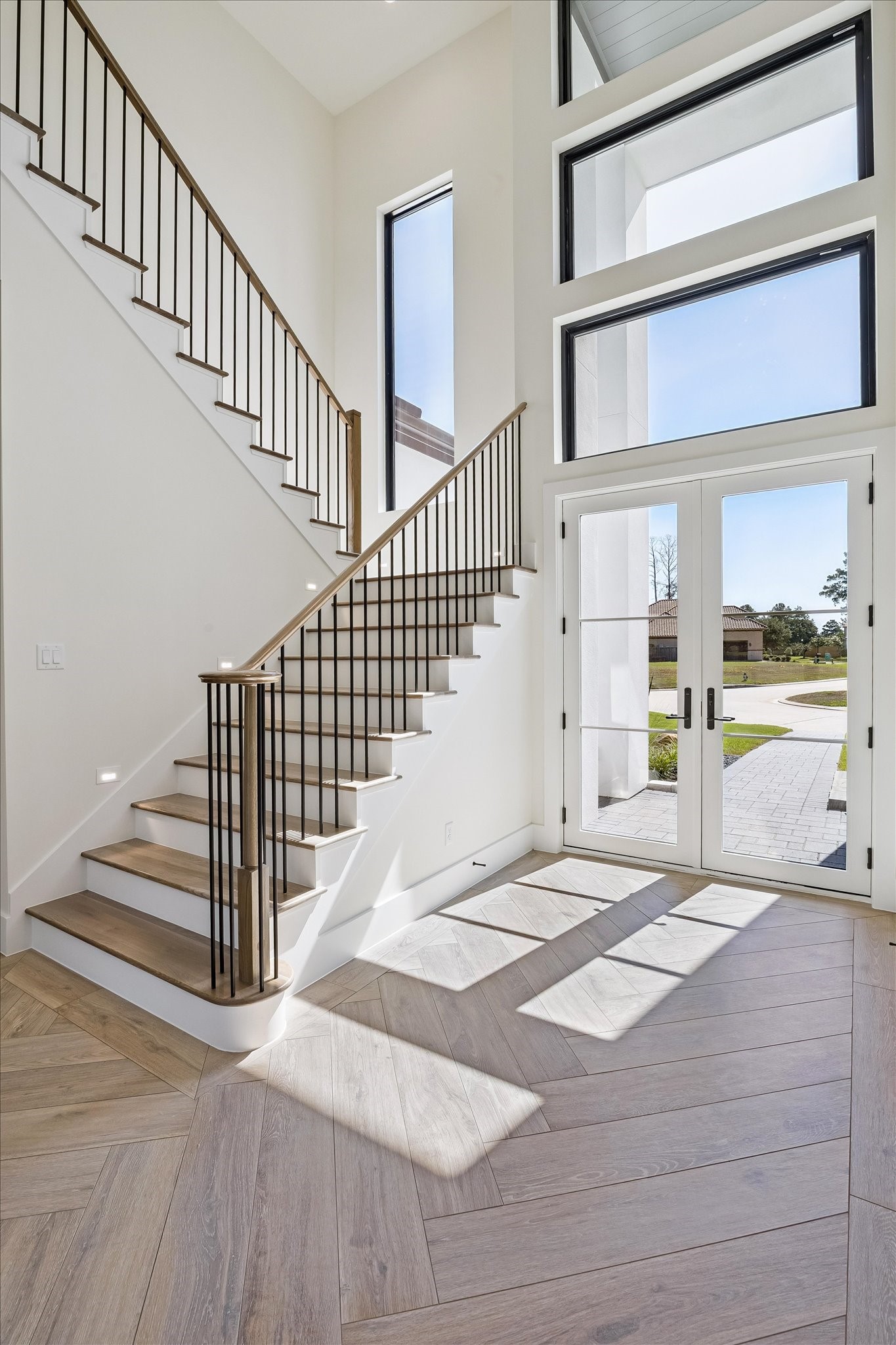 Sun-filled foyer with soaring ceilings, dramatic windows, and a modern staircase over wide-plank flooring.