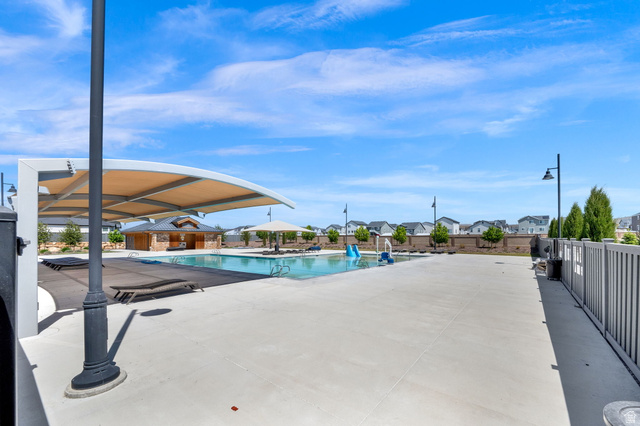 6120 North Sheps Ridge Road Eagle Mountain, UT 84005 - Photo 21 of 43 Community swimming pool featuring a curved shade structure, a separate pool house building with stone detailing, an adjacent splash pad with blue and white water features, and expansive concrete decking