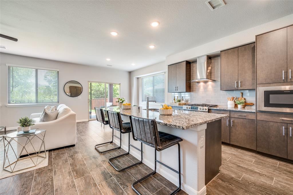 1240 Morning Ridge Trail Aubrey, TX 76227 - Photo 7 of 25 Kitchen featuring backsplash, dark brown cabinets, a breakfast bar, an island with sink, and light stone countertops