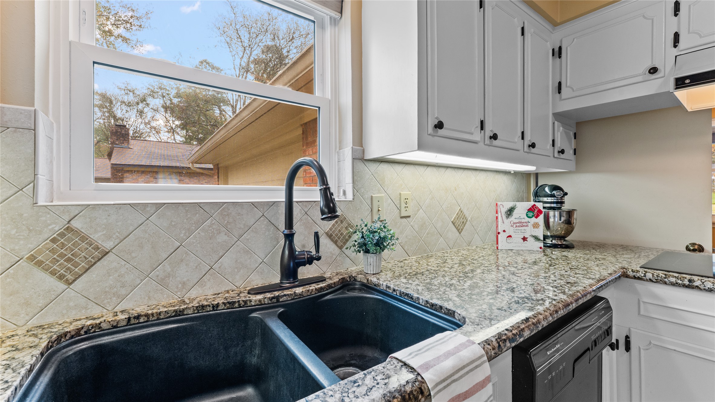 208 Magnolia Way Huntsville, TX 77320 - Photo 10 of 26 This kitchen features white cabinetry, granite countertops, and an updated tile backsplash. It includes a large double sink, electric cooktop, 2-built-in stoves, and dishwasher. Natural light comes in through a window over the sink, and there's an open view to the adjacent dining area with tiled flooring. (Fridge is excluded from sale).