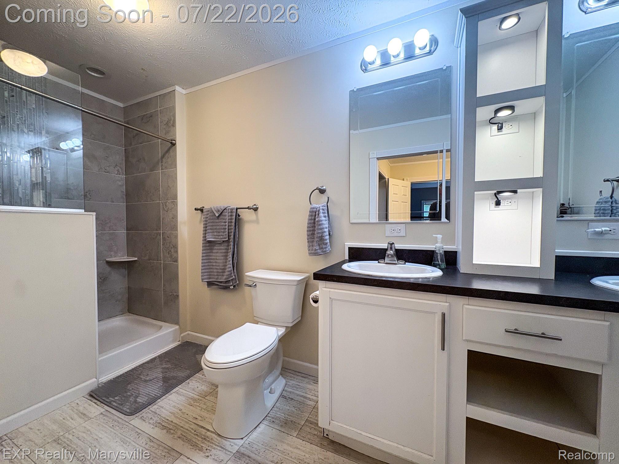 2601 Beach Road Port Huron, MI 48060 - Photo 27 of 34 Bathroom featuring a tiled shower enclosure with a built-in shelf, light wood-finish flooring, a white vanity with a dark countertop and integrated sink, an illuminated recessed shelving unit, and a vanity mirror with overhead lighting