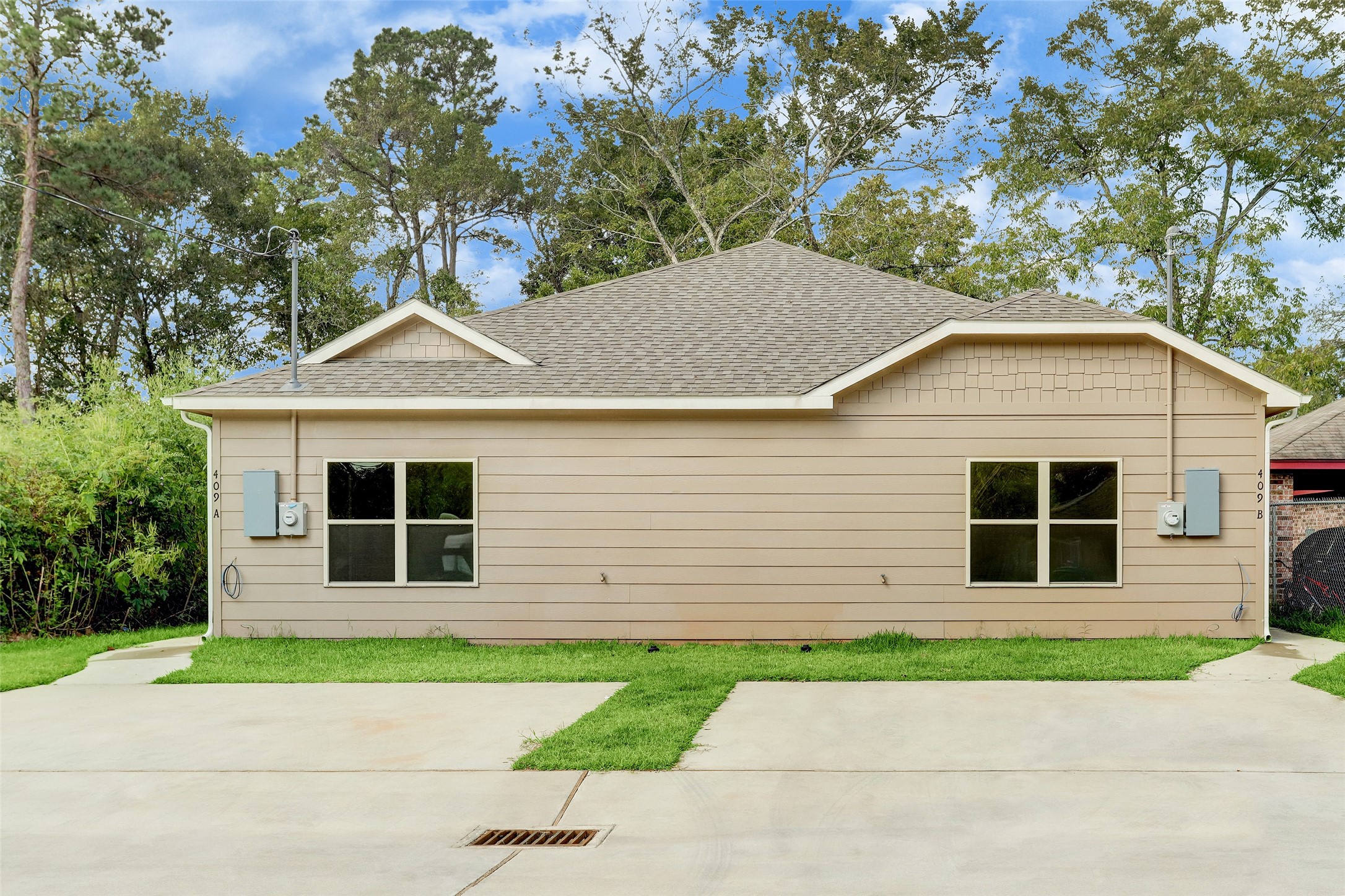 409 South 12th Street, Unit B Conroe, TX 77301 - Photo 2 of 14 This is a rear view of a single-story house with a beige exterior, two windows, and a gabled roof. It features a concrete driveway and is surrounded by trees, offering a private and serene setting.