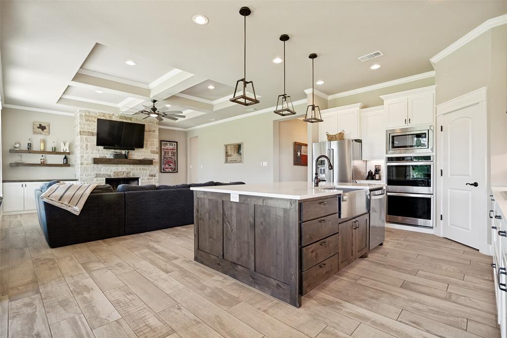 6913 Veal Station Road Weatherford, TX 76085 - Photo 11 of 31 Open-concept kitchen and living area featuring wood-finish flooring, a stone fireplace with a wood mantle, a coffered ceiling with recessed lighting, a kitchen island with a built-in sink and pendant lighting, and stainless steel appliances