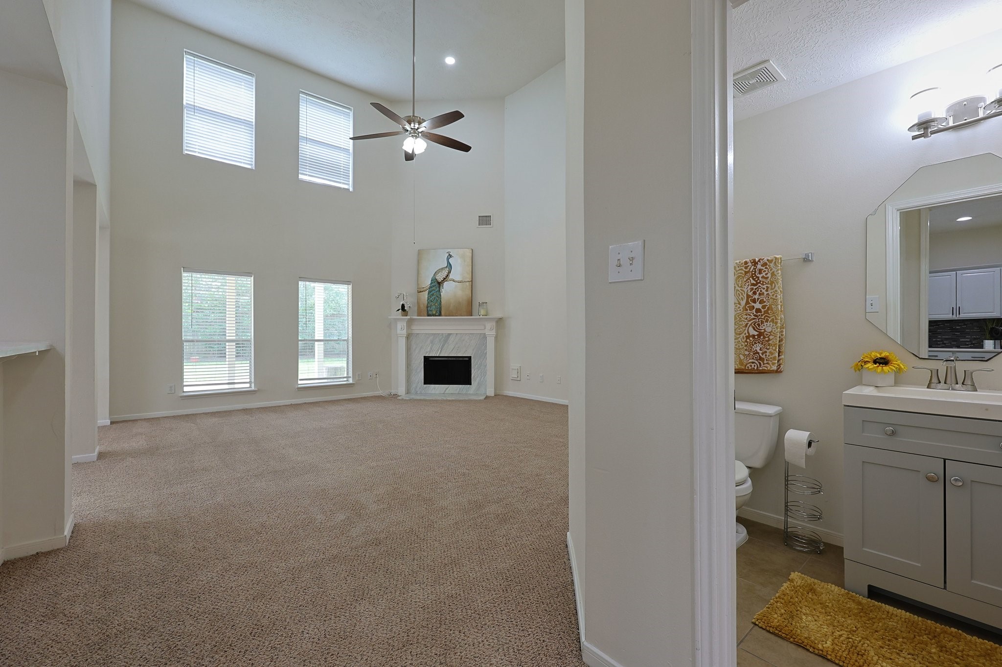 23019 Eastgate Village Drive Spring, TX 77373 - Photo 17 of 46 This photo showcases a spacious living area with high ceilings, large windows, and a ceiling fan, providing ample natural light. The room features a fireplace with decorative artwork above. Adjacent is a bathroom with a modern vanity and bright lighting.