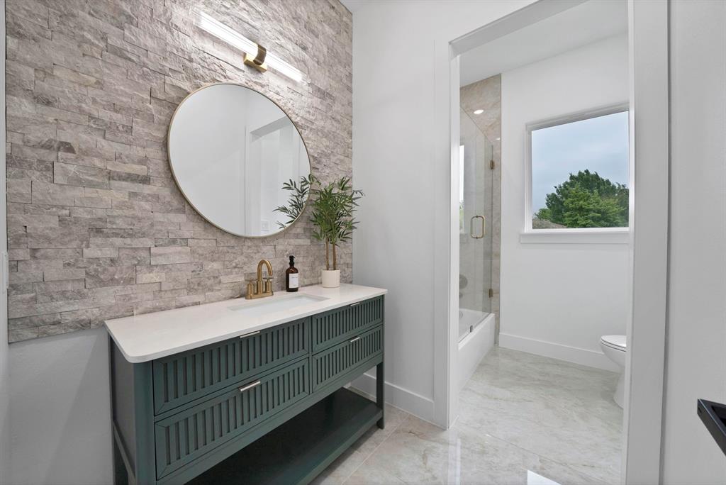 710 Gibson Road Waxahachie, TX 75165 - Photo 27 of 36 Contemporary bathroom featuring a stylish vanity with a white countertop and gold-toned fixtures, a circular mirror, and a textured stone accent wall