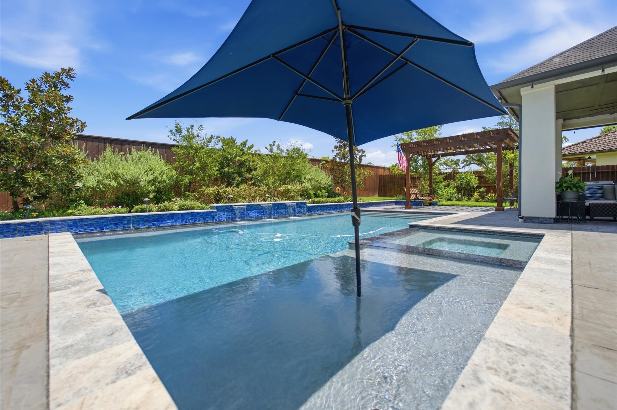 23722 Thortons Park Lane Spring, TX 77389 - Photo 42 of 48 This photo showcases a beautiful backyard with a modern swimming pool, featuring a sunken area with a large umbrella for shade. The pool is surrounded by a stone patio and lush landscaping, creating a relaxing outdoor oasis. A pergola and seating area are visible in the background, enhancing the space for entertaining.