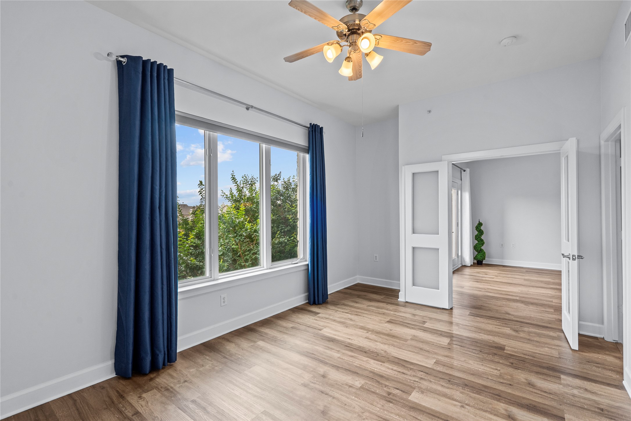 1900 Genesee Street, Unit 203 Houston, TX 77006 - Photo 20 of 30 This bright room features large windows with blue curtains, a ceiling fan, and wood-style flooring. The open doorway leads to another spacious area, enhancing the airy feel.