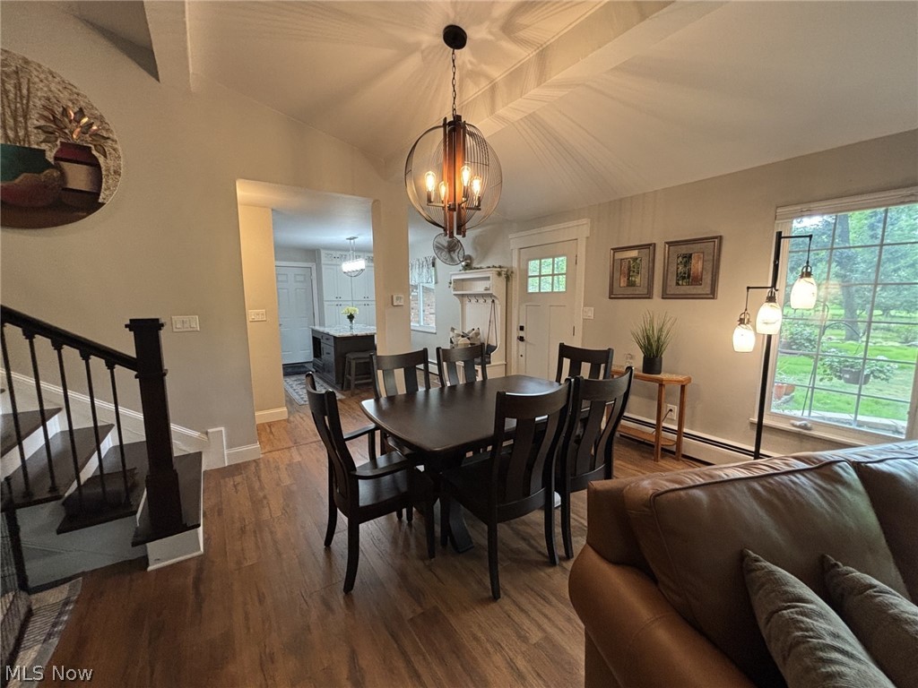 Vaulted ceiling with exposed beams, engineered wood flooring, staircase with dark metal railing, large picture window with custom shades, and a dark stone fireplace hearth.
