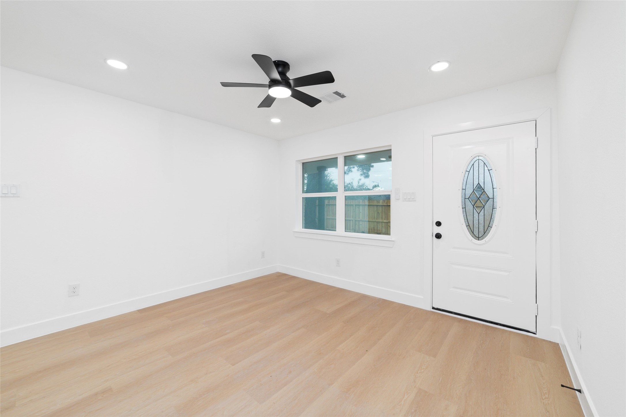 5709 Langley Road Houston, TX 77016 - Photo 6 of 27 Bright, modern room with light wood flooring, white walls, and recessed lighting. Features a ceiling fan and a decorative front door with a window, offering a clean and inviting entryway.